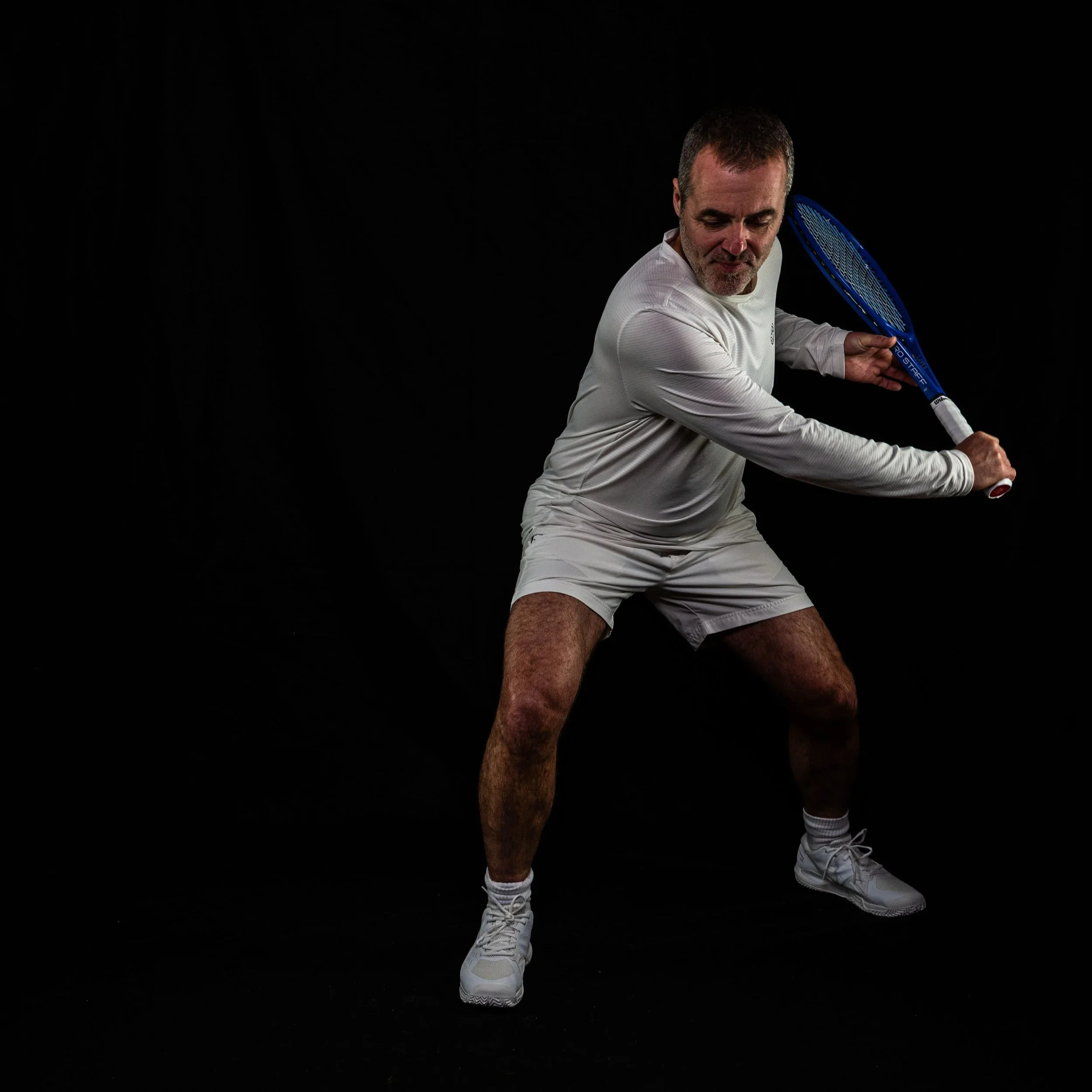 Ein Mann in weißer Sportkleidung mit Tennisschläger in einer Tennis-Haltung vor einem schwarzen Hintergrund.