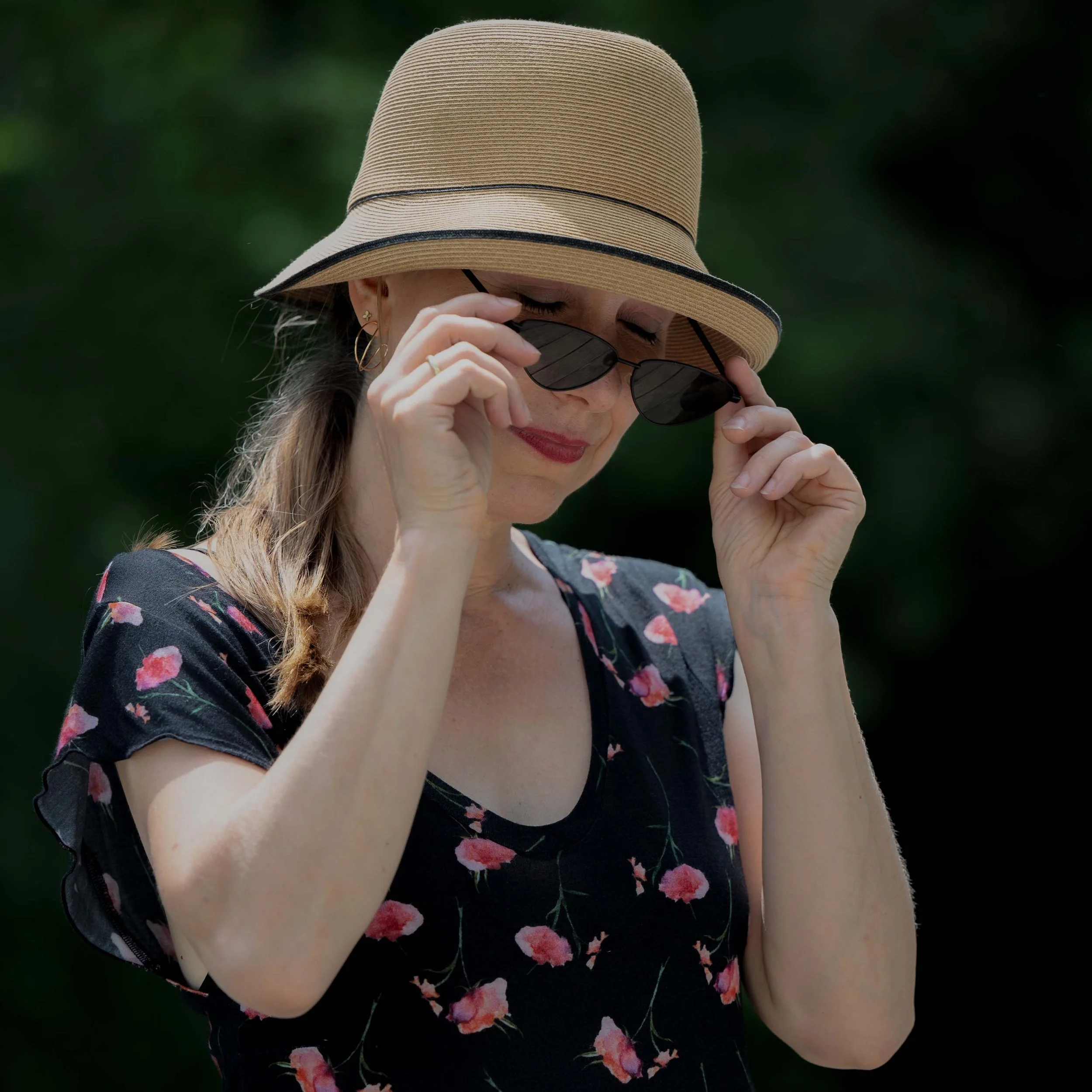 Frau trägt Sommerhut und Sonnenbrille, trägt ein schwarzes Kleid mit rosafarbenen Blumen, blickt nach unten, im Hintergrund grüne Natur.
