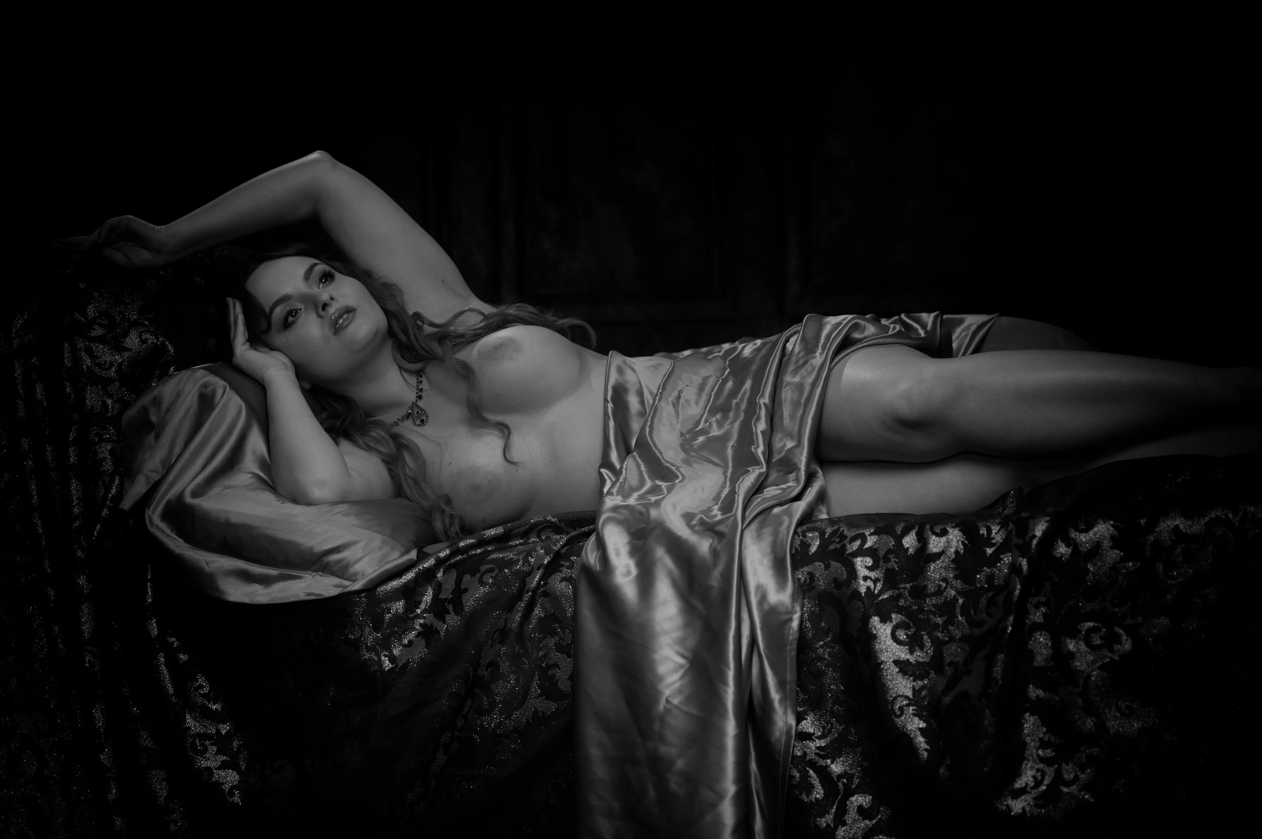 Eine Frau liegt nackt auf einem Bett, umgeben von seidenen Stoffen, in einem dunklen Raum, schwarz-weiße Fotografie