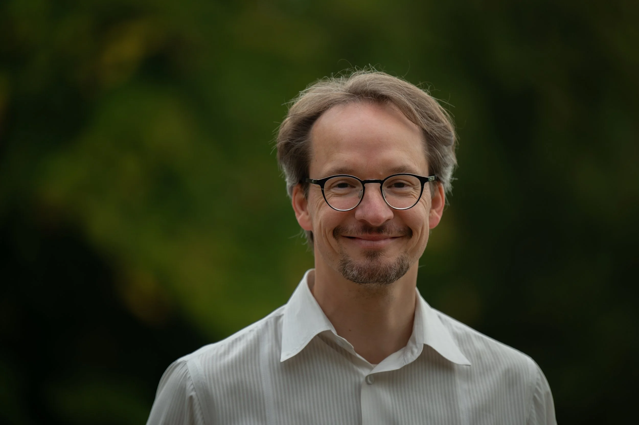 Ein Mann mit Brille, dunklem Haar und Bart trägt ein weißes Hemd und lächelt vor einem unscharfen grünen Hintergrund.