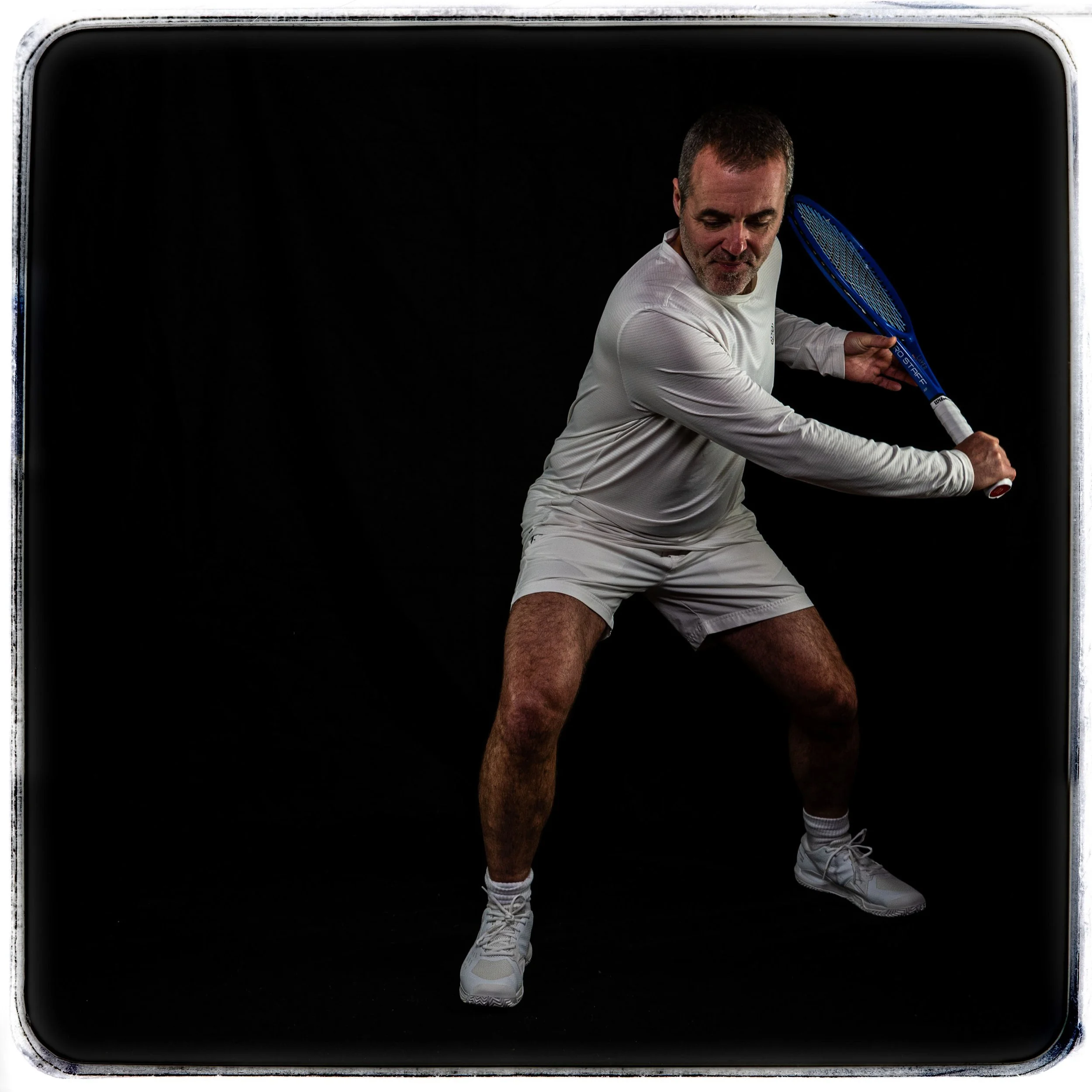 Mann in weißer Sportbekleidung hält einen Tennisracket und ist in einer Tennisposition, vor schwarzem Hintergrund.