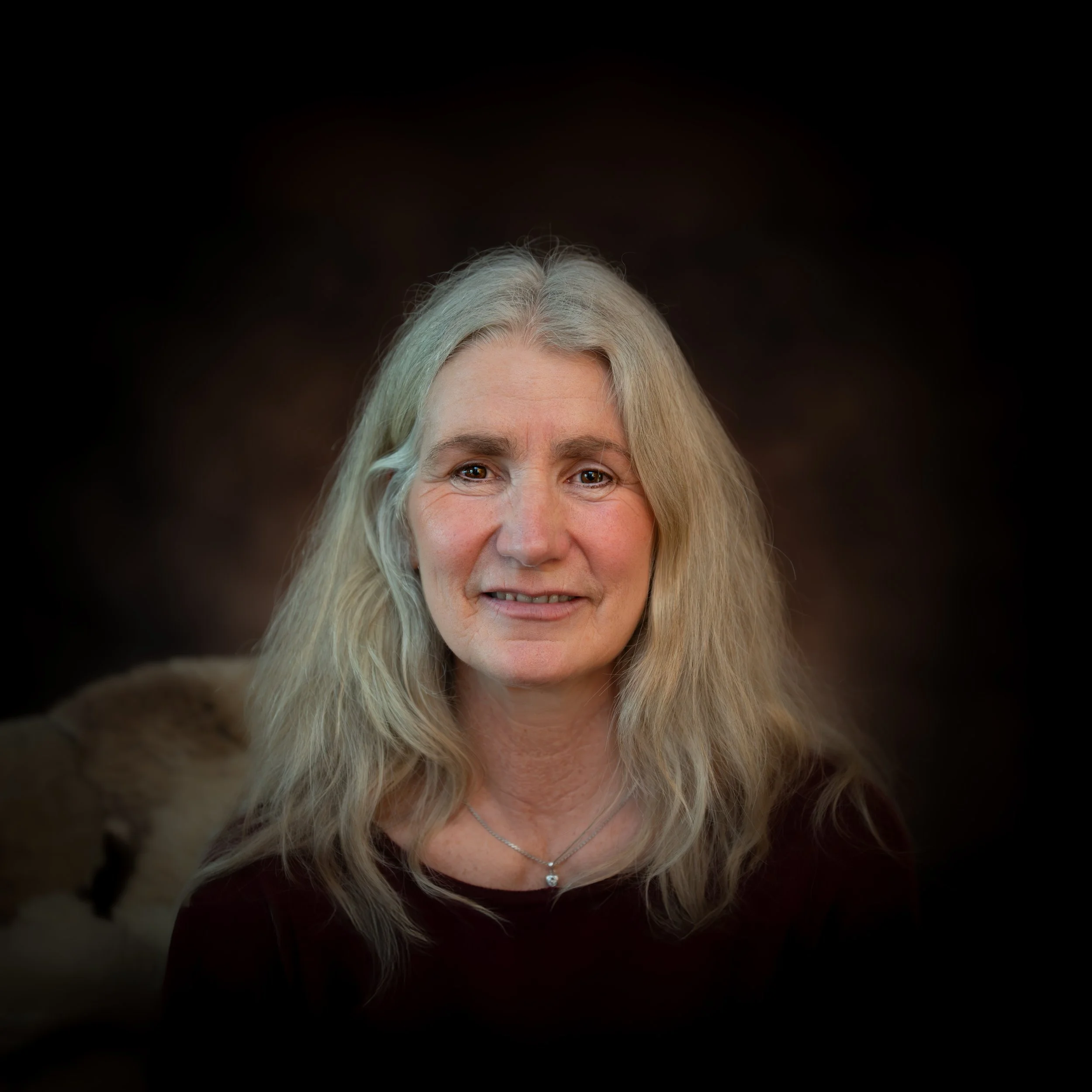 Porträt einer älteren Frau mit langen grauen Haaren, die lächelt, vor dunklem Hintergrund.