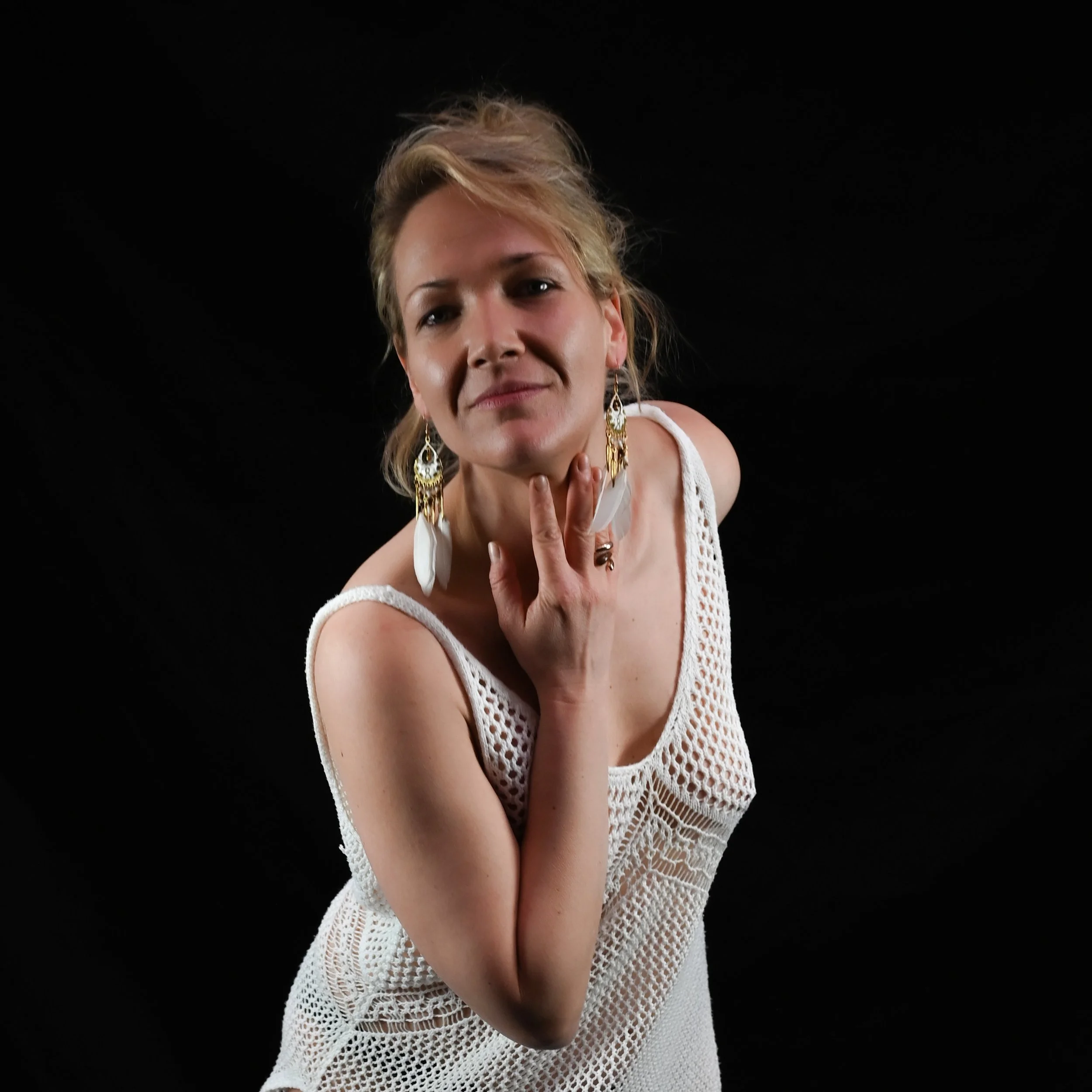Nahaufnahme einer jungen Frau mit lockigem, hellbraunem Haar vor schwarzem Hintergrund, trägt große goldene und weiße Ohrringe sowie ein weißes, offenporiges Kleid.