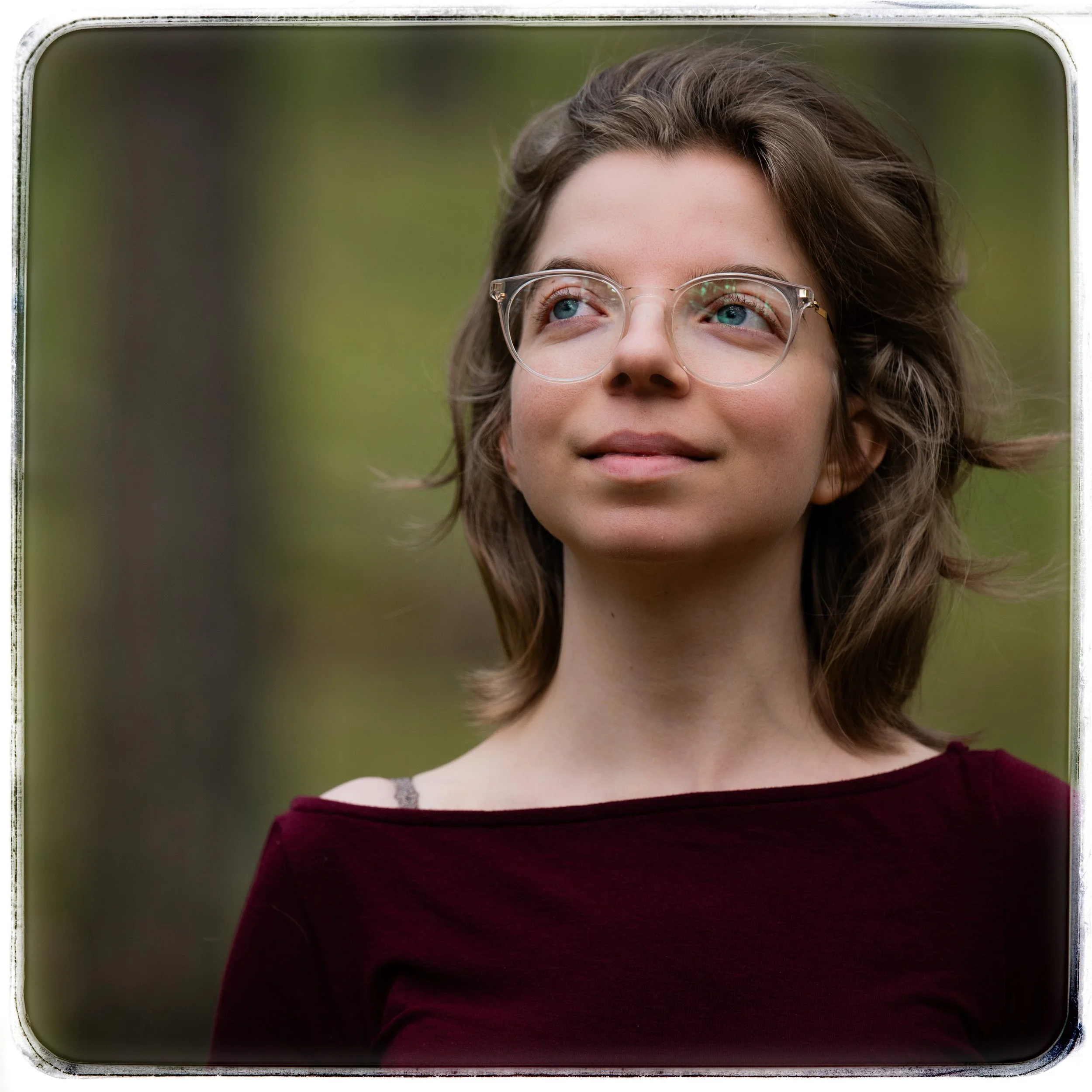 Junge Frau mit Brille im Freien, lächelnd, grüner Wald im Hintergrund.
