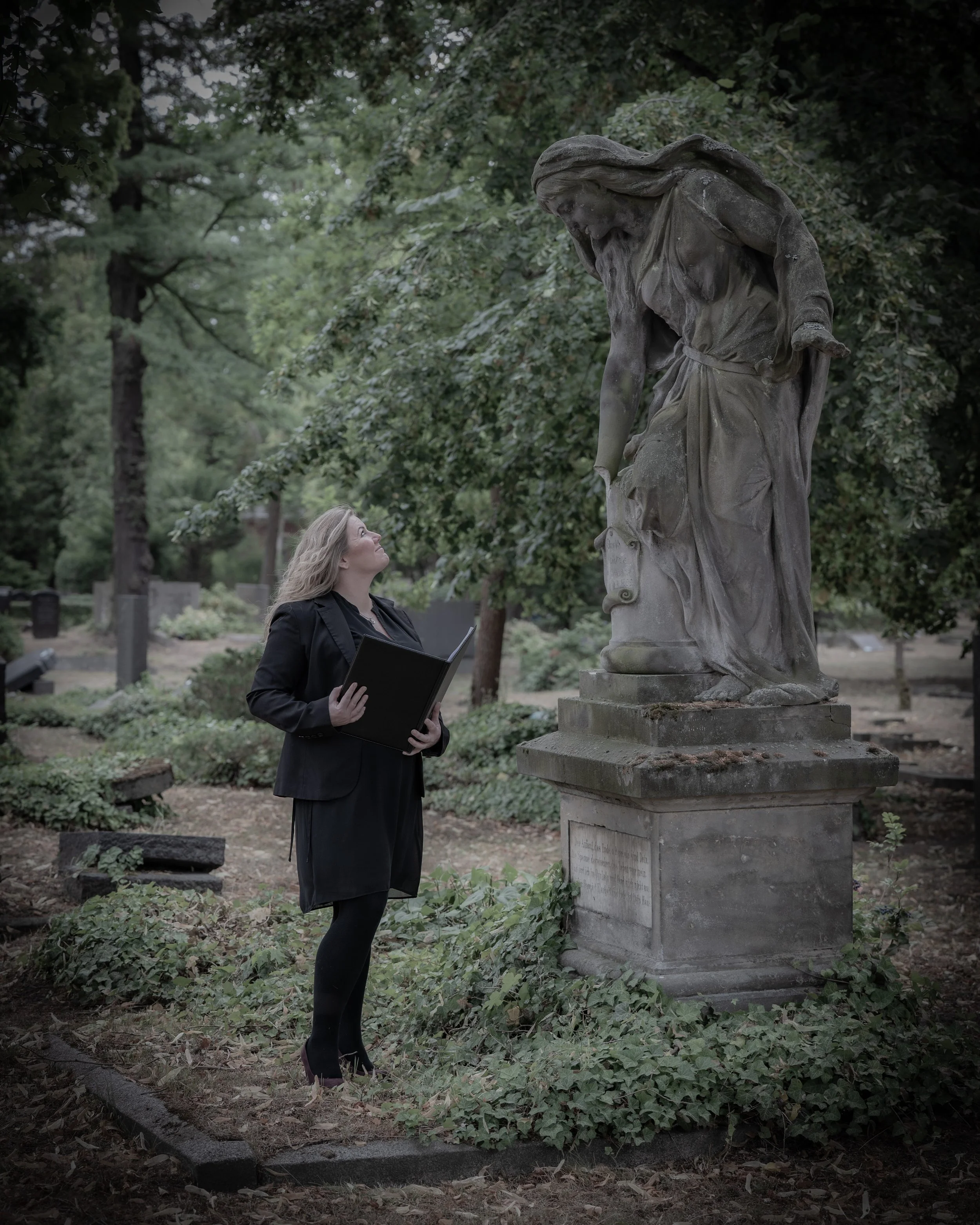Eine Frau in formeller Kleidung steht vor einer Statue in einem Park und liest aus einem schwarzen Buch.
