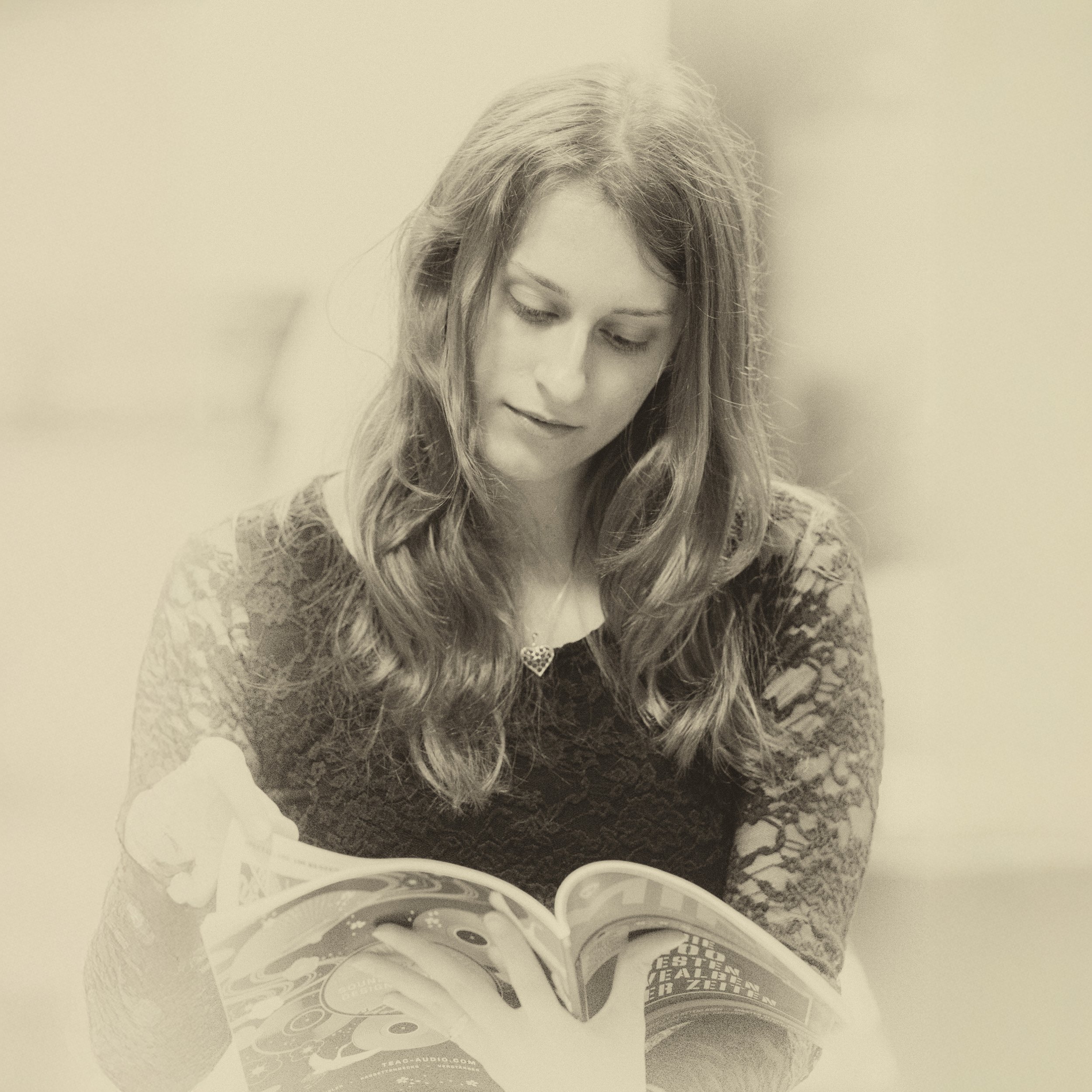 Junge Frau mit langen, lockigen Haaren liest eine Zeitschrift, trägt ein Top mit Spitze und ein Halsband mit Herzanhänger, sepiafarbener Ton.