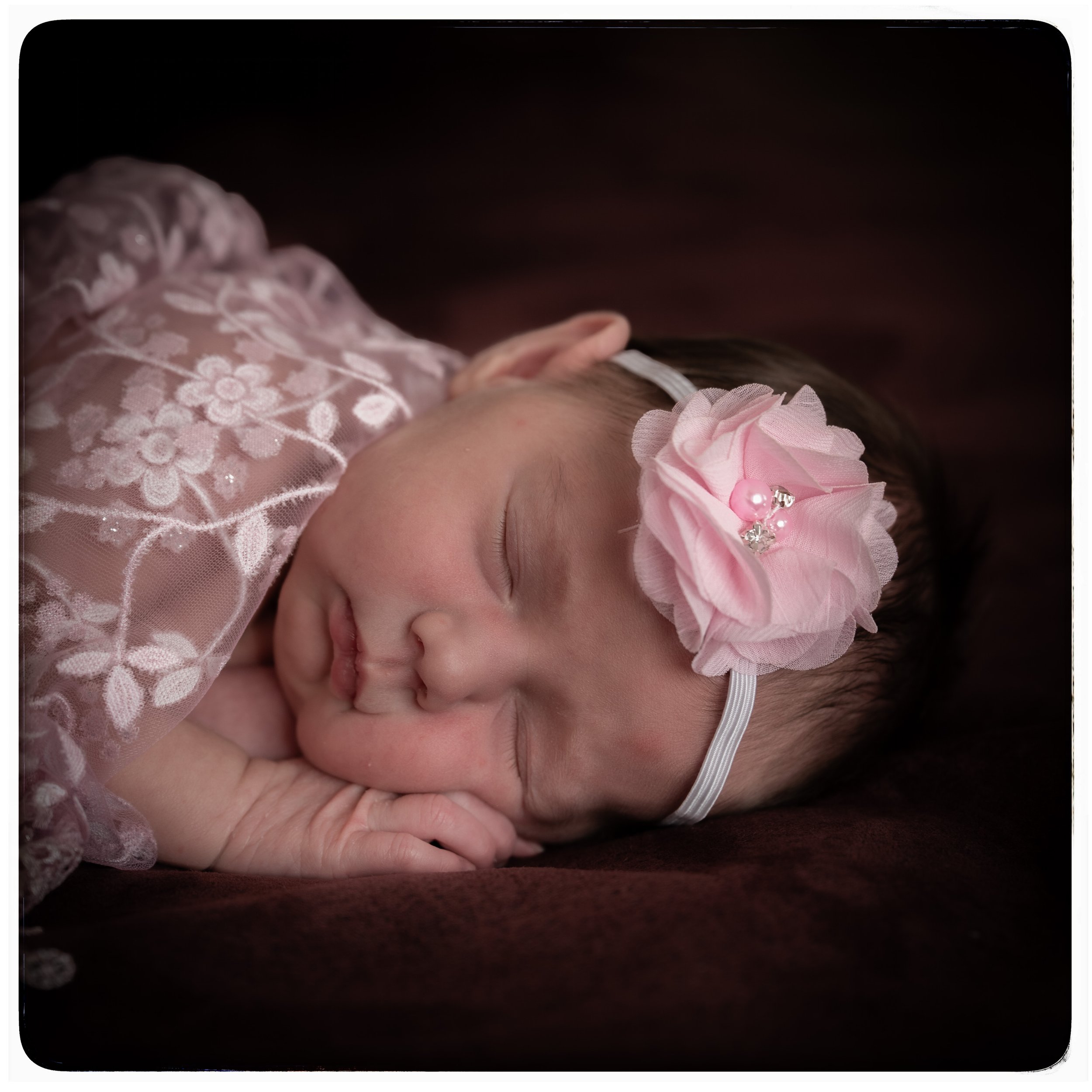 Kleines schlafendes Baby mit rosa Blumen Stirnband, in rosa Spitze Kleidung, auf braunem Hintergrund.