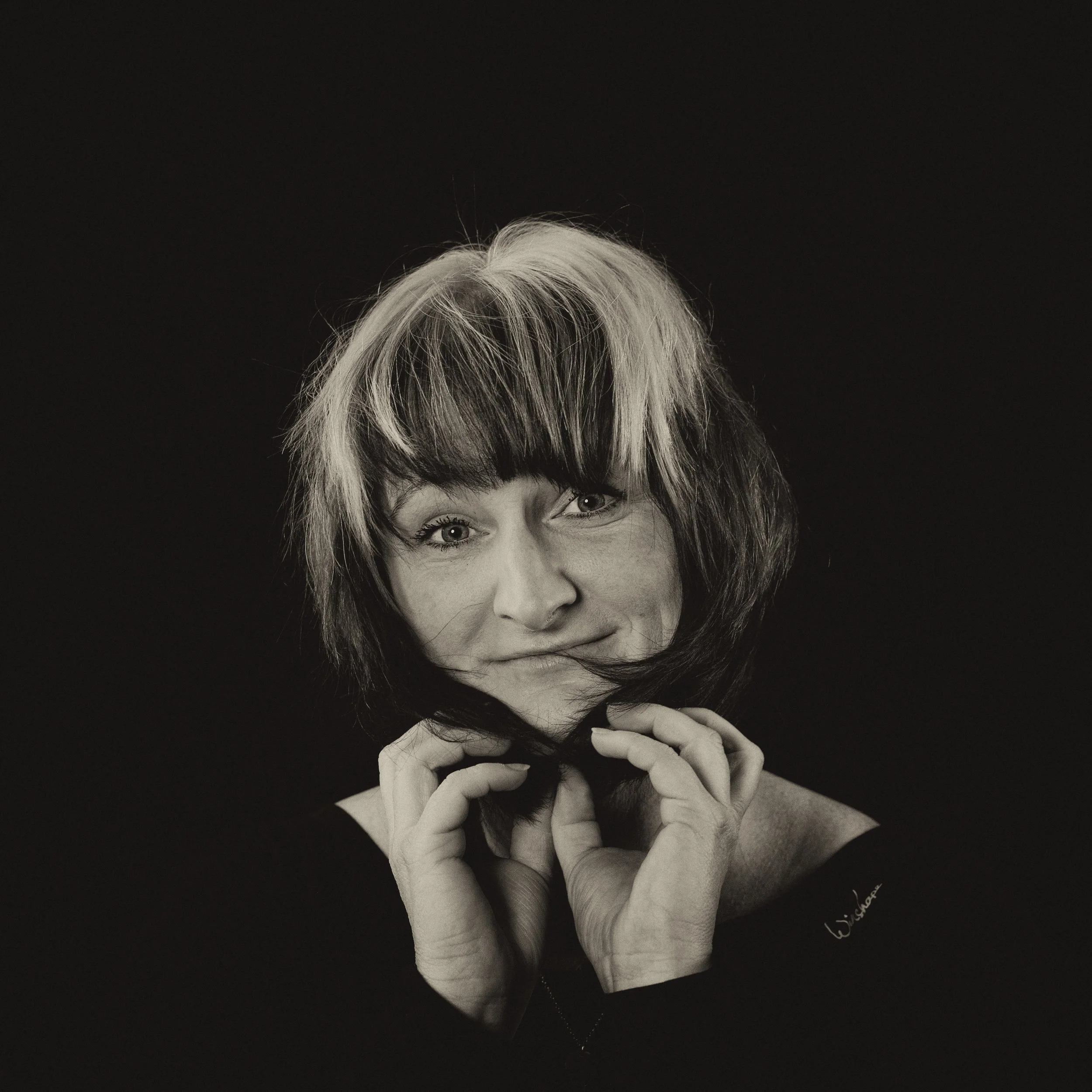 Schwarz-weiß Portrait einer Frau mit mixed Haarfarbe, die eine Haarlocke in den Händen hält und direkt in die Kamera schaut.