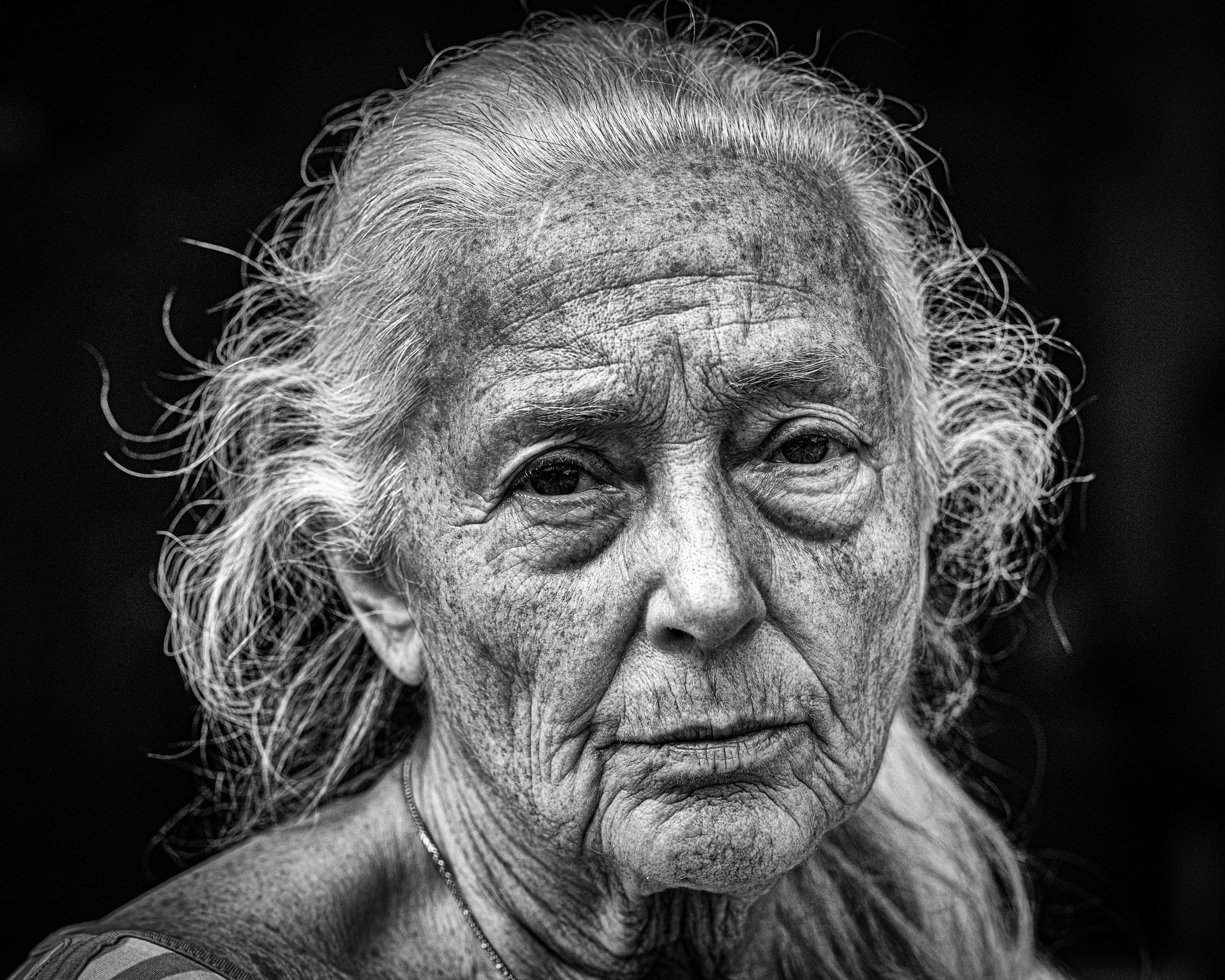 Schwarz-Weiß-Porträt einer alten Frau mit langen, grauen, lockigen Haaren, sichtbaren Falten und einem ernsten Ausdruck.