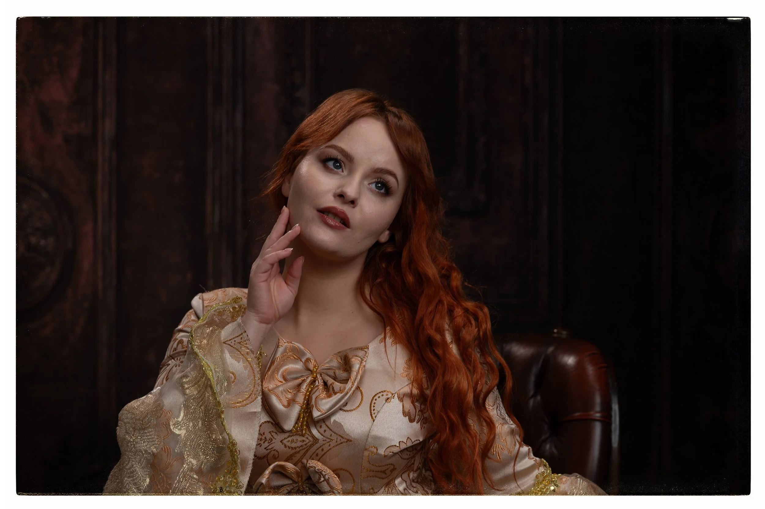 Eine Frau mit langen roten Locken, die auf einem braunen Ledersessel sitzt, trägt ein elegantes, dekoratives Kleid mit Goldakzenten und einem großen Schleifenkragen, vor einem dunklen Holzwand-Hintergrund.