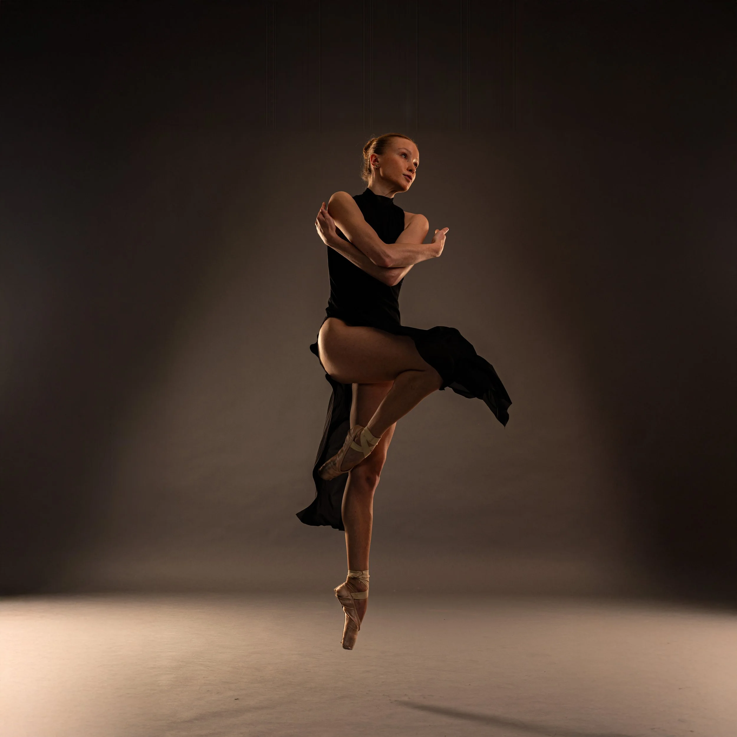Balletttänzerin in einer schwarzen Kleidung, die auf Zehenspitzen steht und eine Pose mit verschränkten Armen einnimmt, auf einer dunklen Bühne