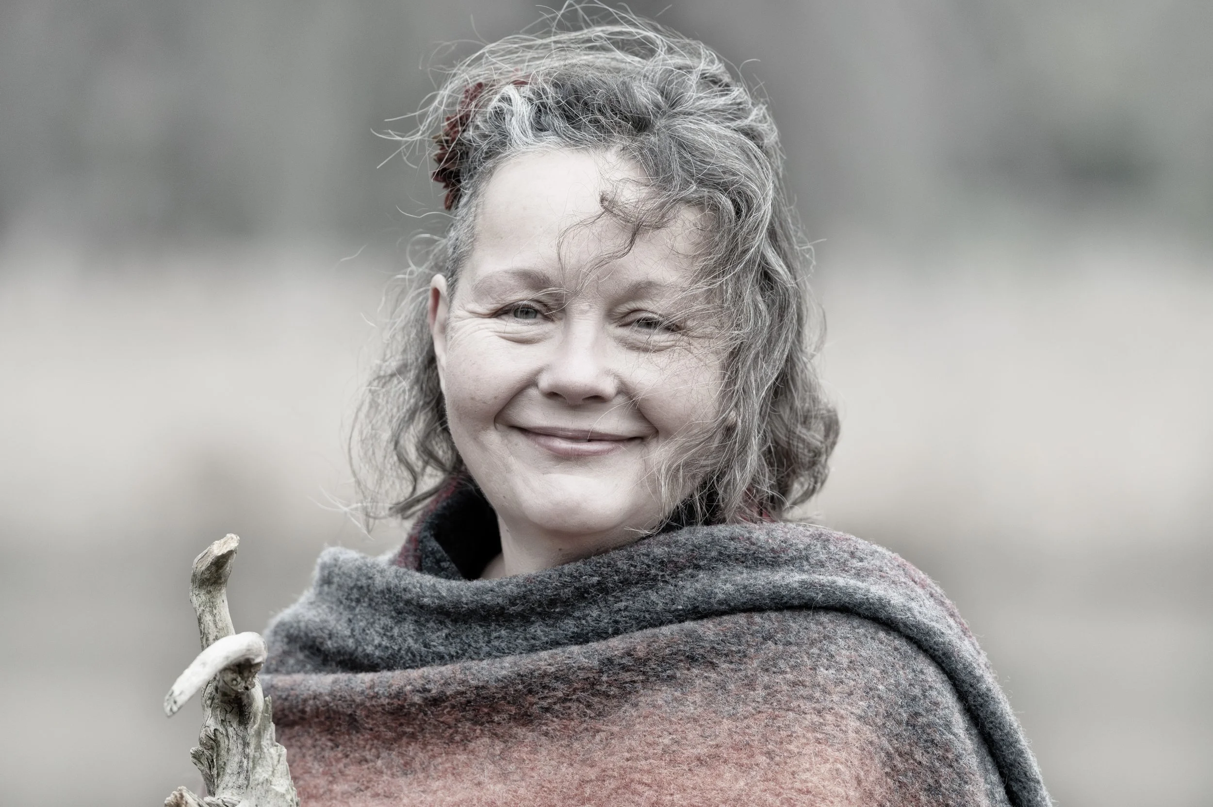 Ein lachender, älterer Frau, die ein bunt gestreiftes Wolltuch trägt, im Freien, hält ein Stück Holz in der Hand.