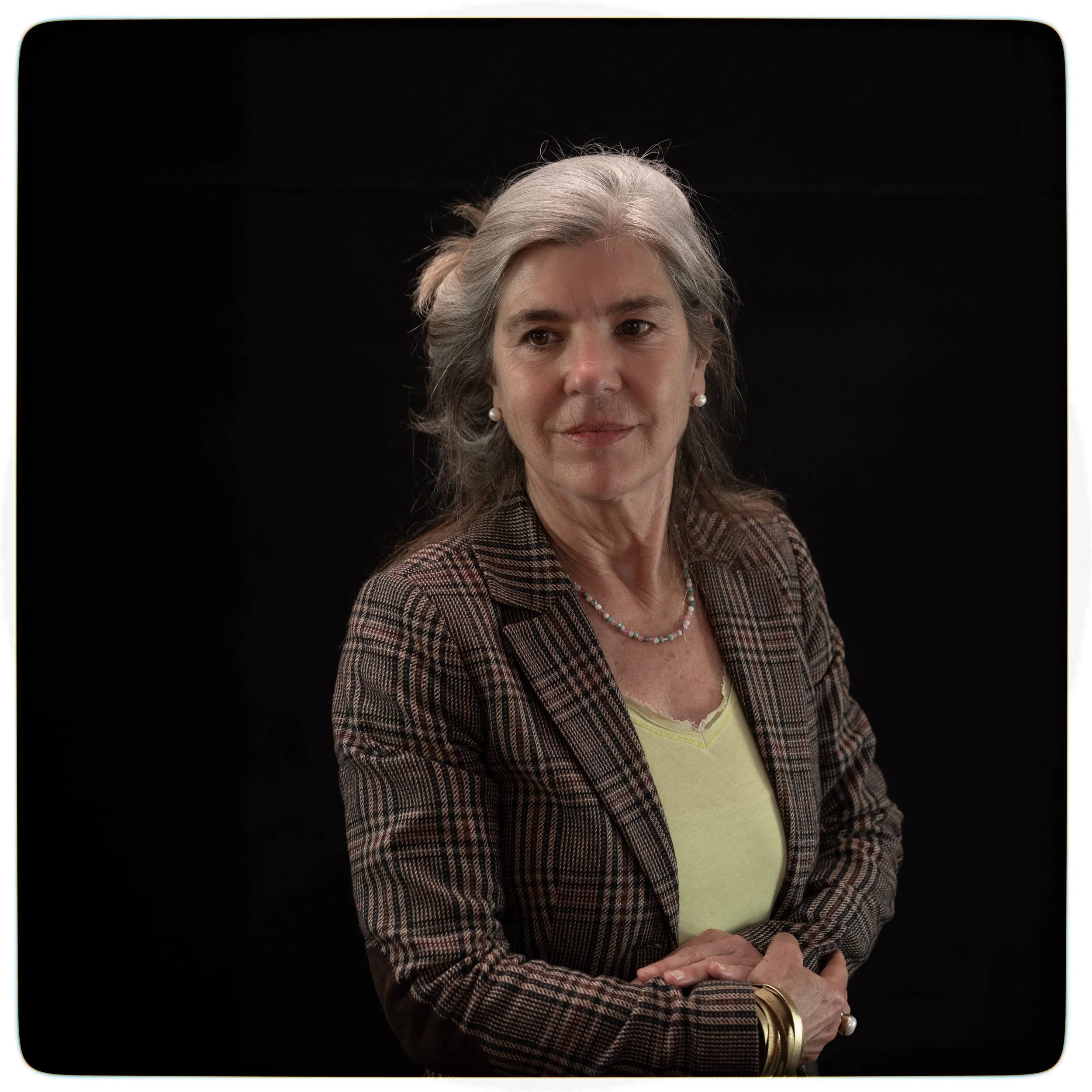 Porträt einer älteren Frau mit grauem Haar vor schwarzem Hintergrund, trägt eine karierte Jacke, gelbes Top, Perlenkette und Armbänder.