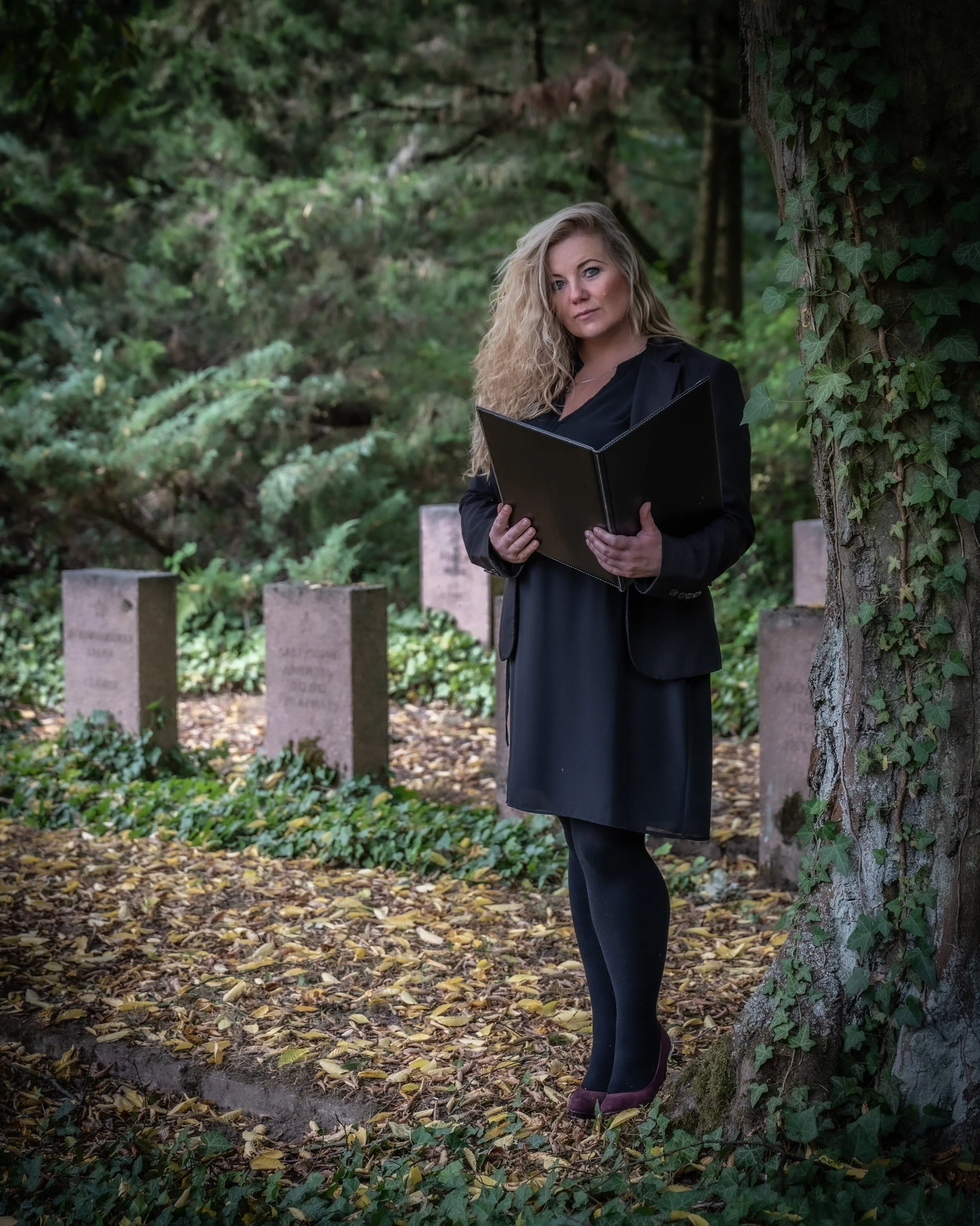 Eine Frau mit lockigem blonden Haar, die in einem schwarzen Kleid und Blazer in einem grünen, bewaldeten Ort steht und ein schwarzes Buch hält.