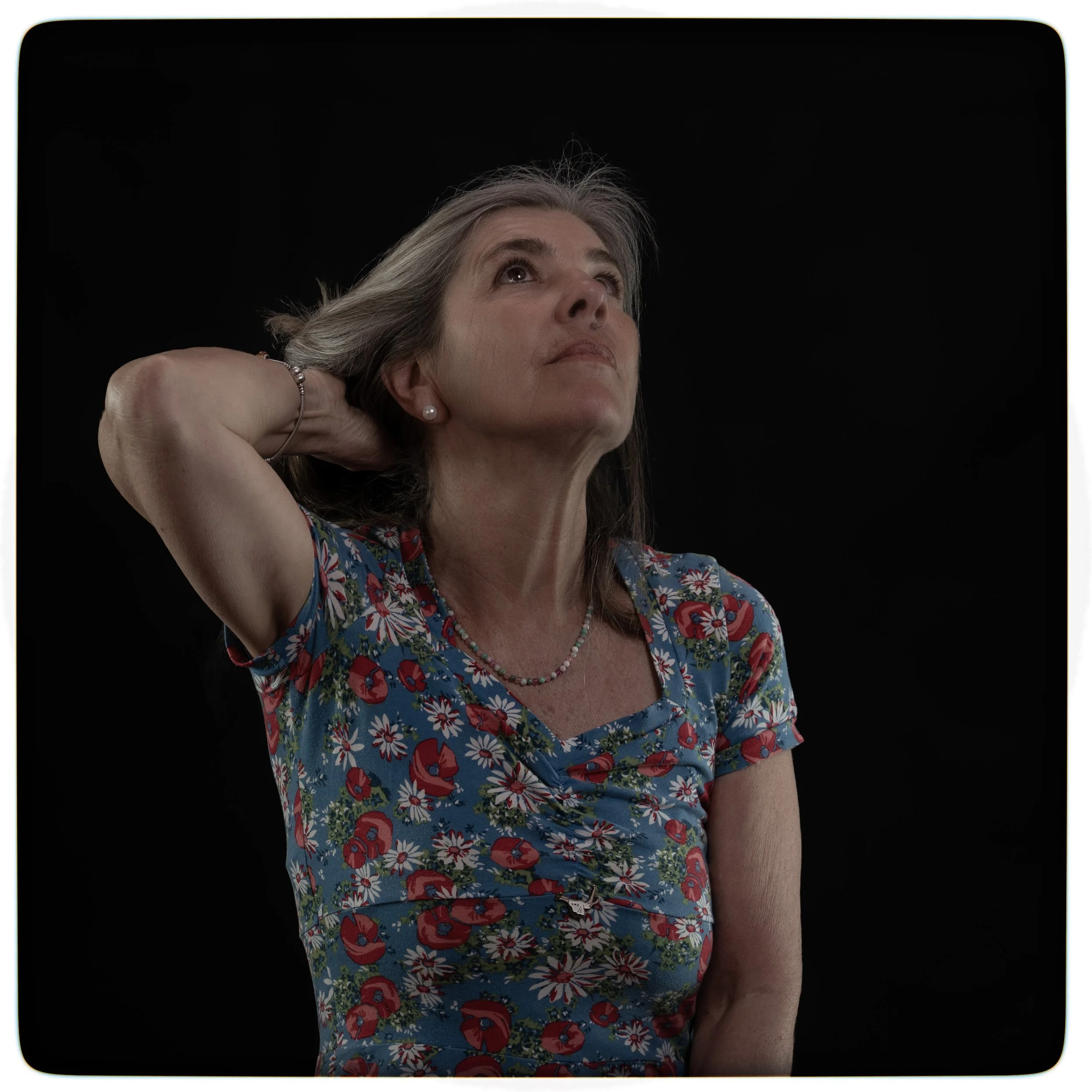 Eine ältere Frau mit grauem Haar, trägt ein blumenmusteriertes Kleid und eine Perlenkette, sieht nach oben und legt eine Hand hinter den Kopf, vor schwarzem Hintergrund.