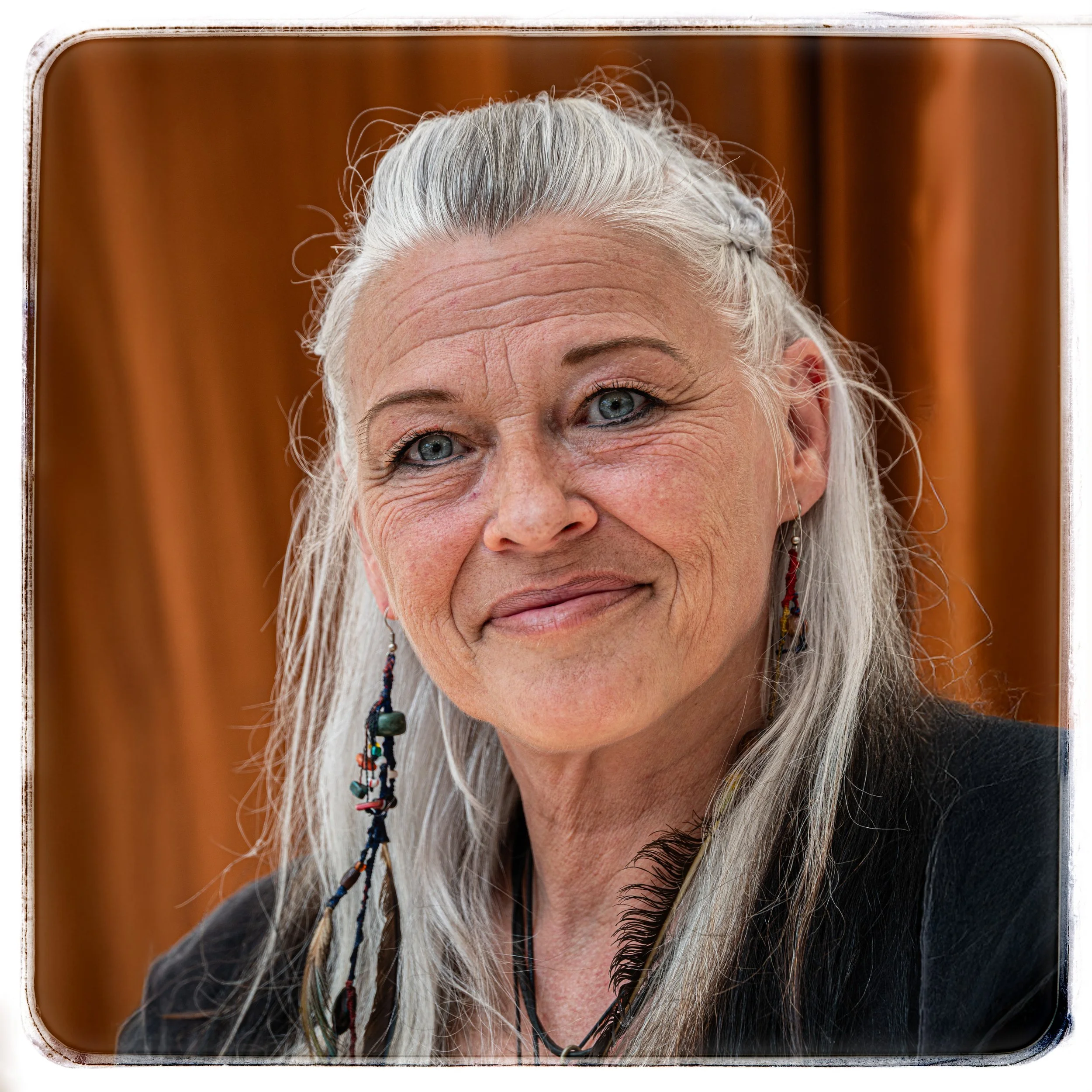 Nahaufnahme eines älteren Mädchens mit langem, grauem Haar, blauen Augen und einem freundlichen Lächeln, tragend bunte, lange Ohrringe, vor einem braunen Stoffhintergrund.