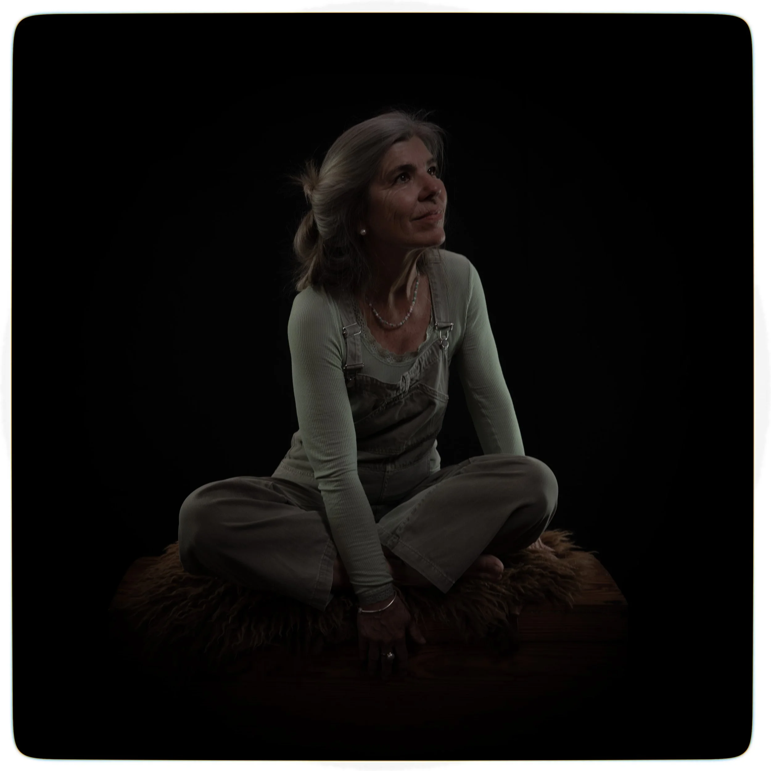 Eine ältere Frau sitzt cross-legged auf einem Pelz auf einem dunklen Hintergrund, trägt einen hellen Pullover und graue Latzhosen, schaut nach oben und lächelt leicht.