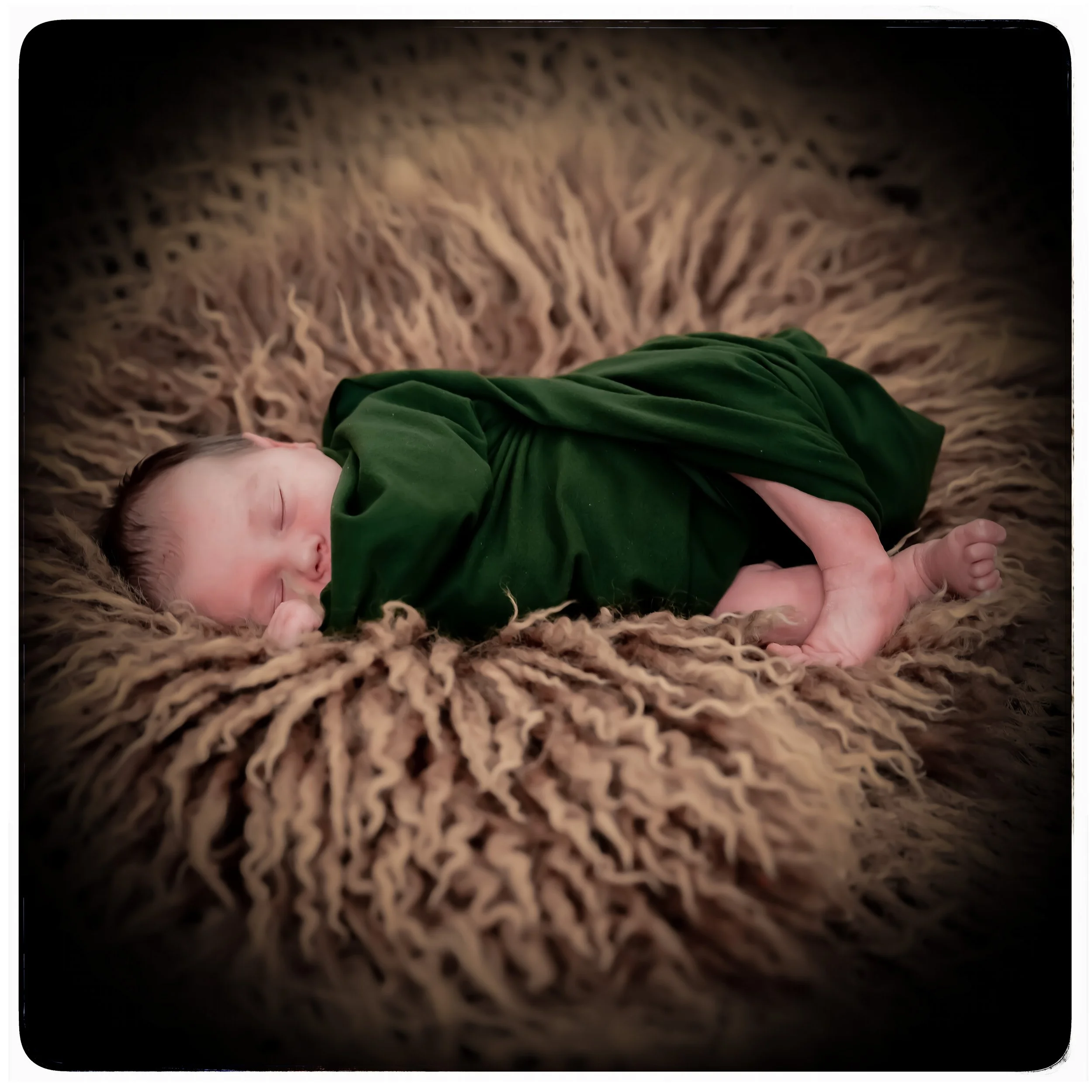 Kleiner schlafender Säugling, eingewickelt in grüne Decke, ruht auf braunem, fluffigem Untergrund.