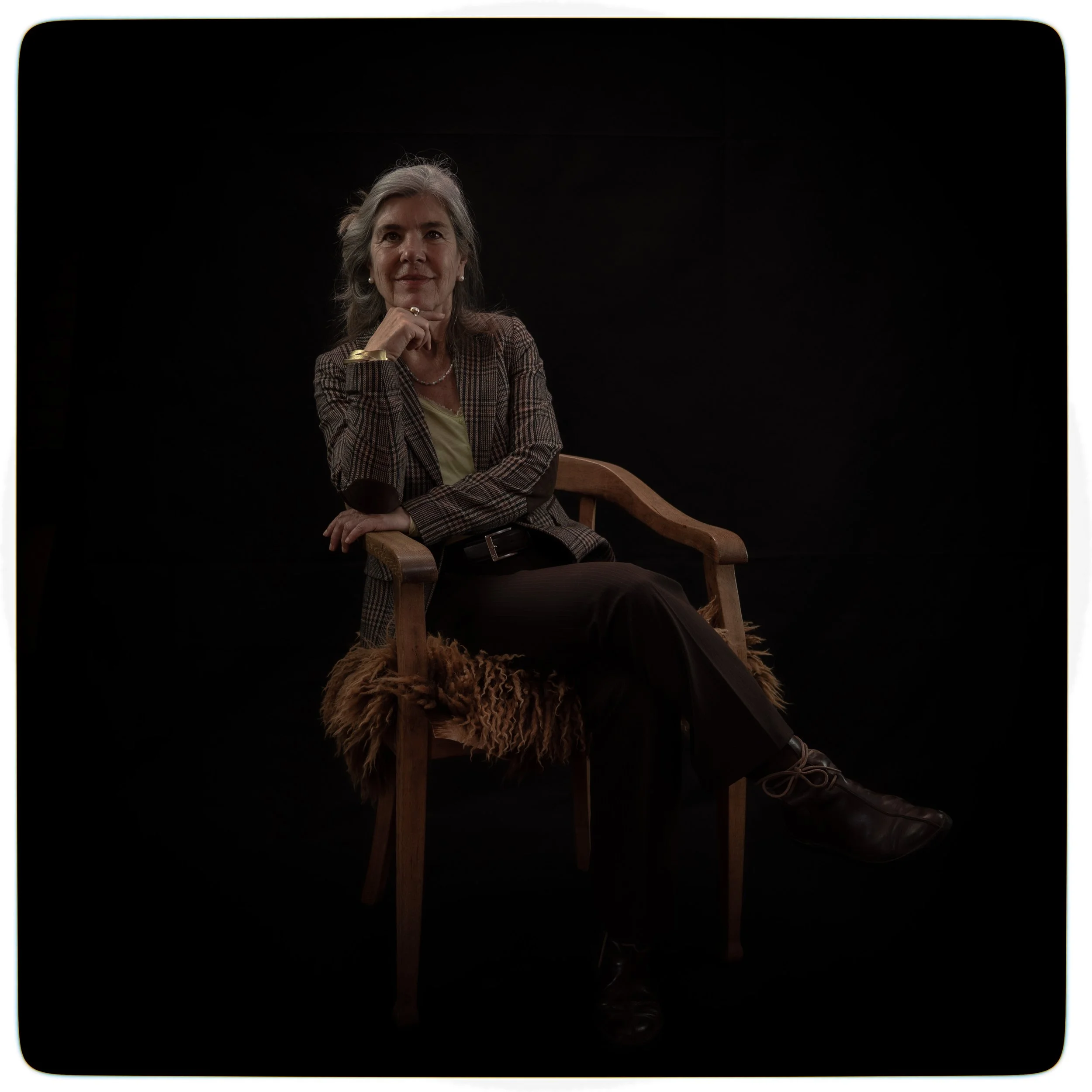 Eine ältere Frau sitzt auf einem Holzstuhl vor einem schwarzen Hintergrund, trägt einen karierten Blazer, eine hellgelbe Bluse, schwarze Hosen und dunkle Schuhe, posiert selbstbewusst mit einer Hand am Kinn.