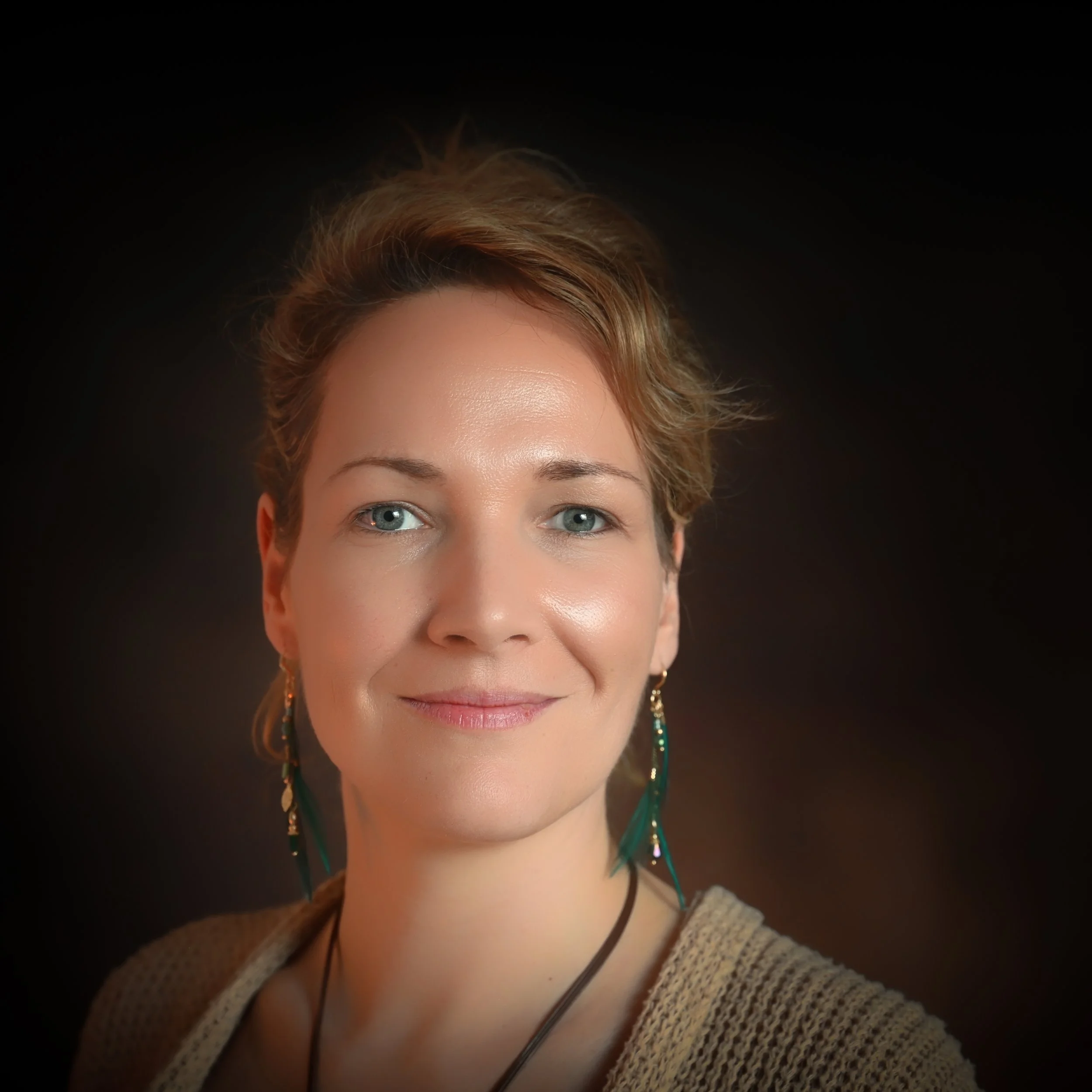 Porträt einer Frau mit blauen Augen, kurzen braunen Haaren und Ohrringen, vor dunklem Hintergrund.