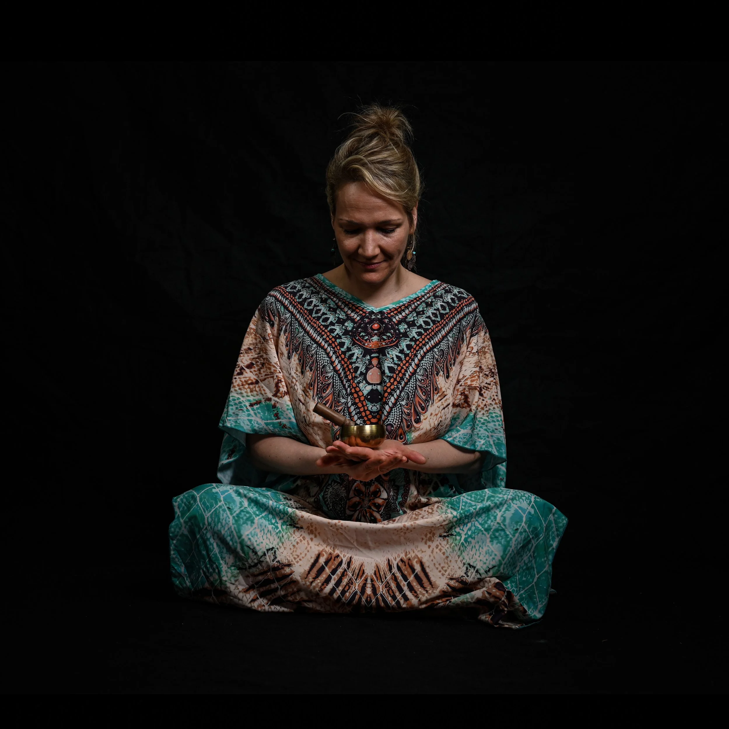 Frau in buntem Kleid sitzt im Schneidersitz vor schwarzem Hintergrund, hält eine kleine Metallglocke in ihren Händen und blickt darauf.