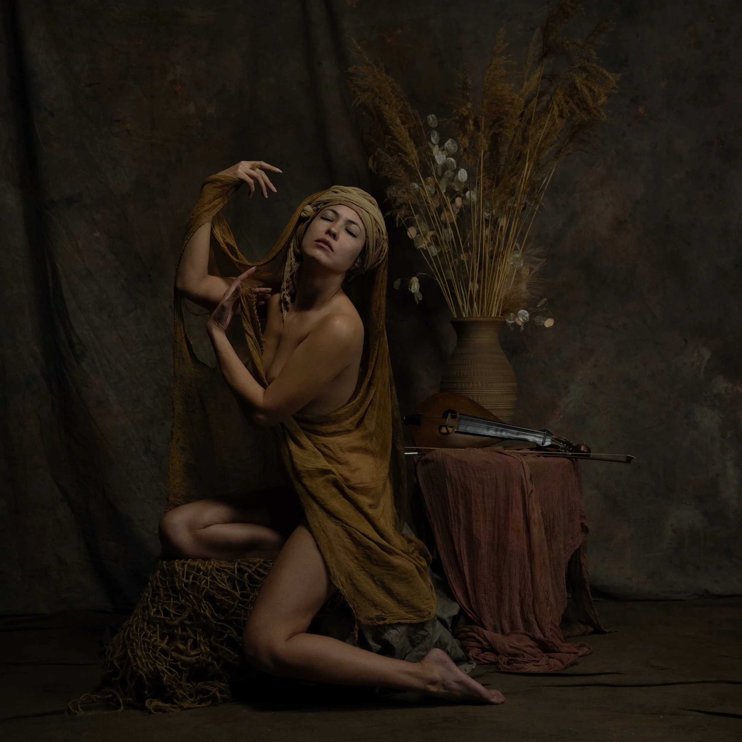Eine Frau posiert nackt vor einem dunklen Hintergrund, eingewickelt in braunes Tuch, mit einer Turban aus Stoff auf dem Kopf. Im Hintergrund steht eine große Vase mit getrockneten Pflanzen, und auf einem Tisch liegen eine Violine und ein Bogen.