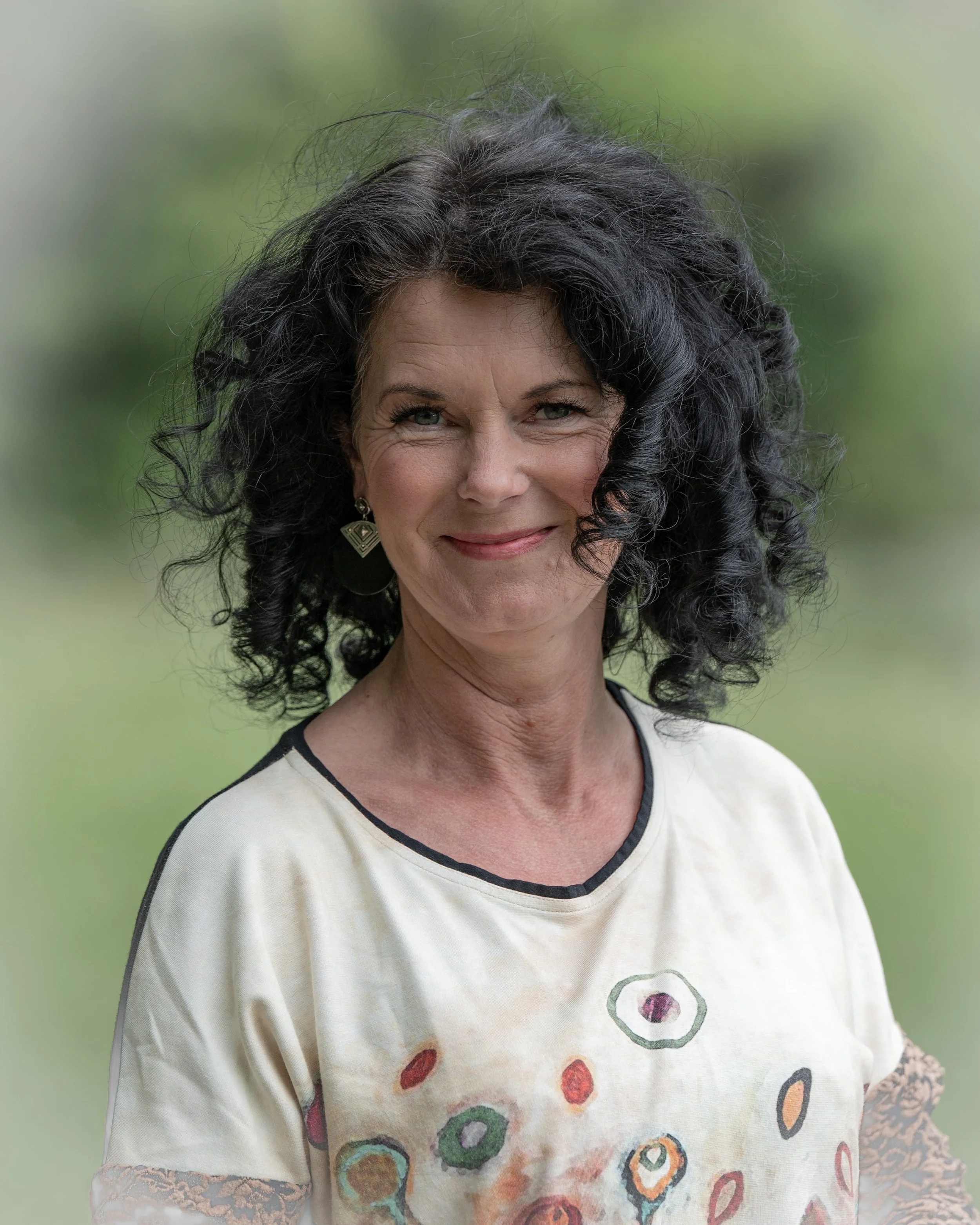 Nahaufnahme einer lachenden Frau mit schwarzen, lockigen Haaren und einem weißen T-Shirt mit bunten Kreisdesigns, im Hintergrund unscharfer grüner Naturbereich