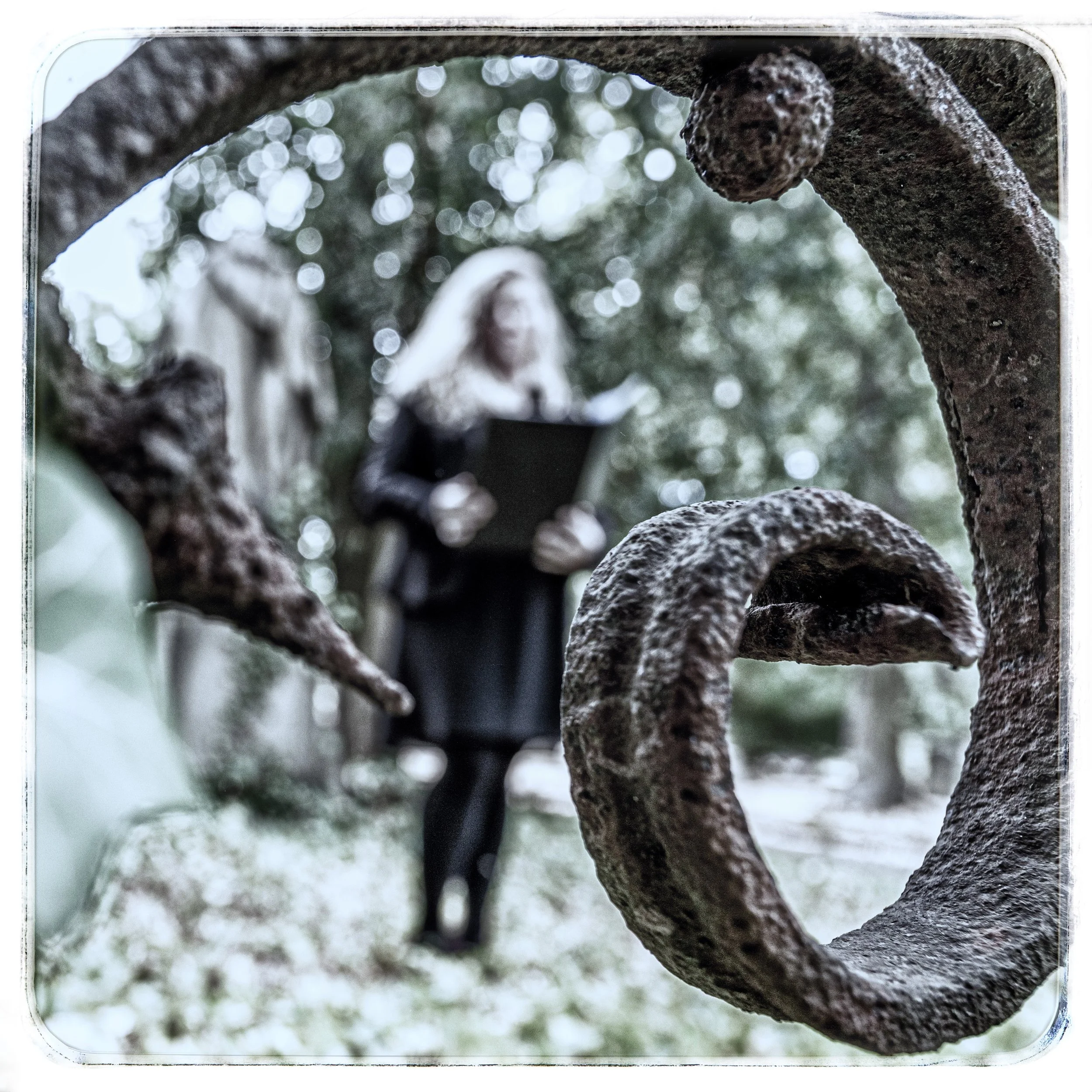 Ein verrosteter Metallring, mit einer rustikalen, gewundenen Oberfläche, im Vordergrund. Im Hintergrund steht eine Frau mit langen blonden Haaren, die ein schwarzes Kleid trägt, in einem Garten mit Bäumen, und liest aus einem schwarzen Buch.