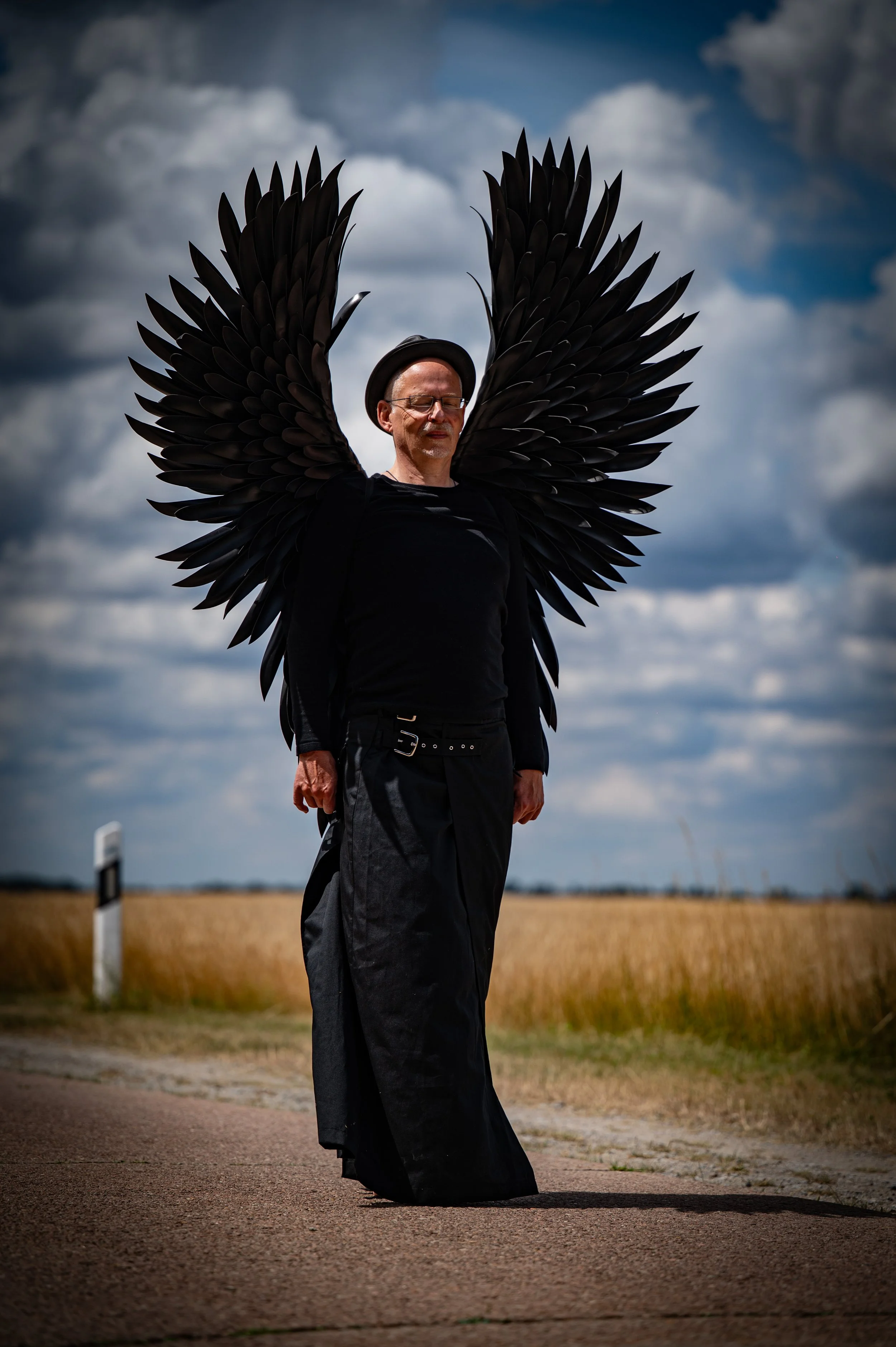 Ein Mann steht auf einer Landstraße mit schwarzen Flügeln, die an seinem Rücken befestigt sind, unter einem bewölkten Himmel.