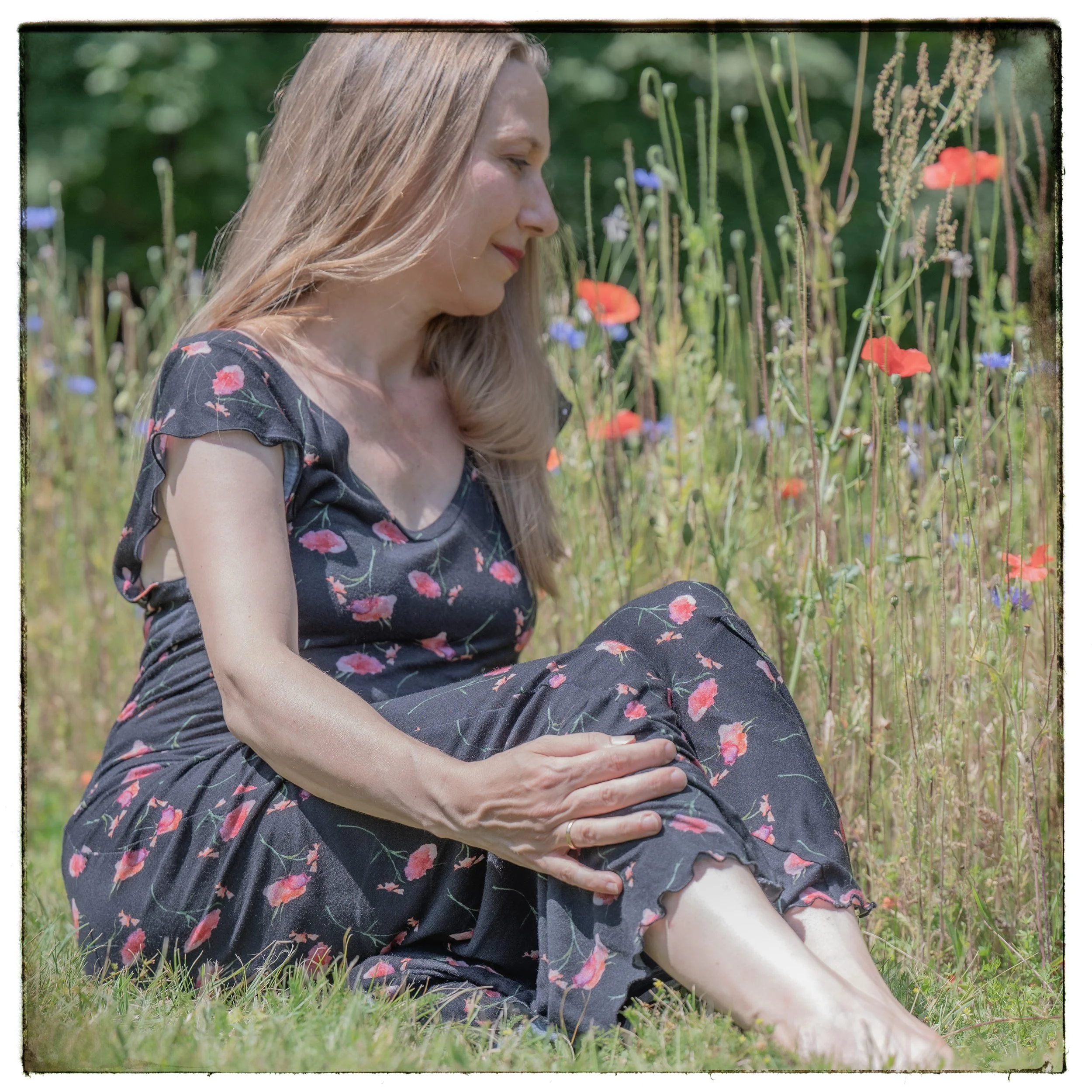 Frau sitzt barfuß im Garten neben bunten Blumen, schaut entspannt nach unten.