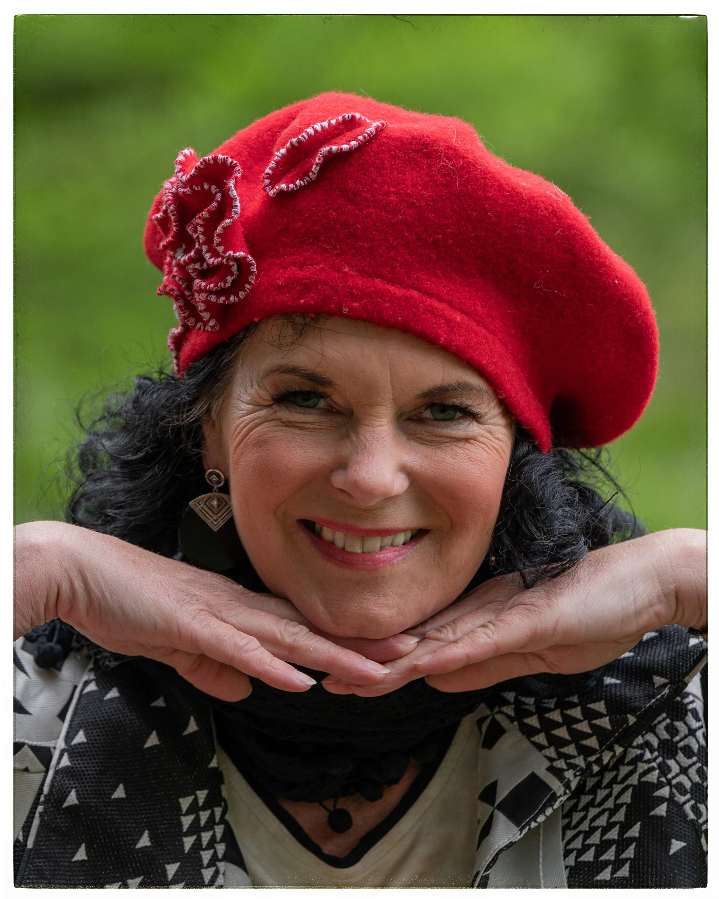 Eine lächelnde Frau mit schwarzen, lockigen Haaren trägt einen roten Hut mit Blumenapplikationen, hält ihr Gesicht mit beiden Händen darunter und trägt ein schwarzes und weißes Muster auf ihrer Kleidung, vor grünem Hintergrund.