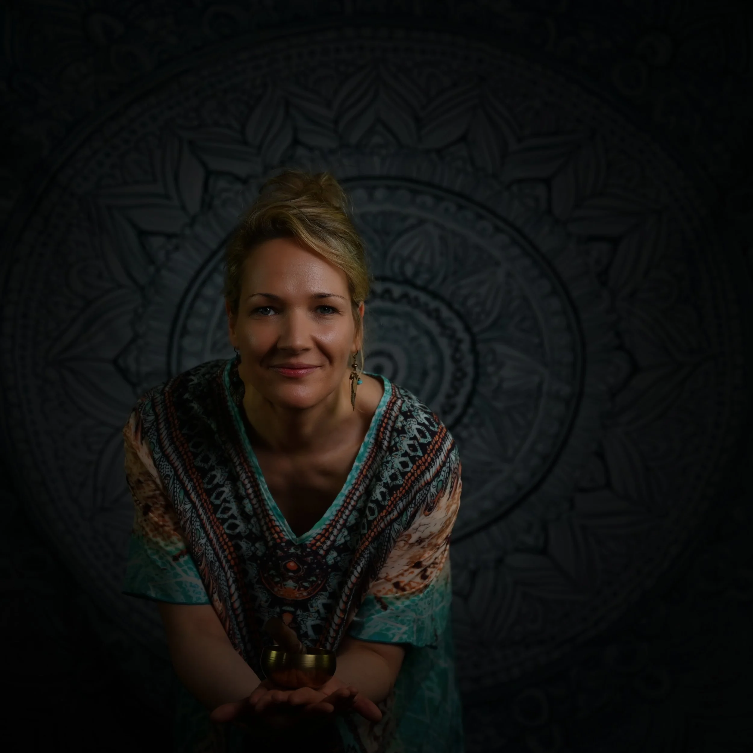 Eine lachende Frau mit lockigem Haar in buntem Sarong hält eine Klangschale in den Händen vor einem mandalaartigen Wandbild.