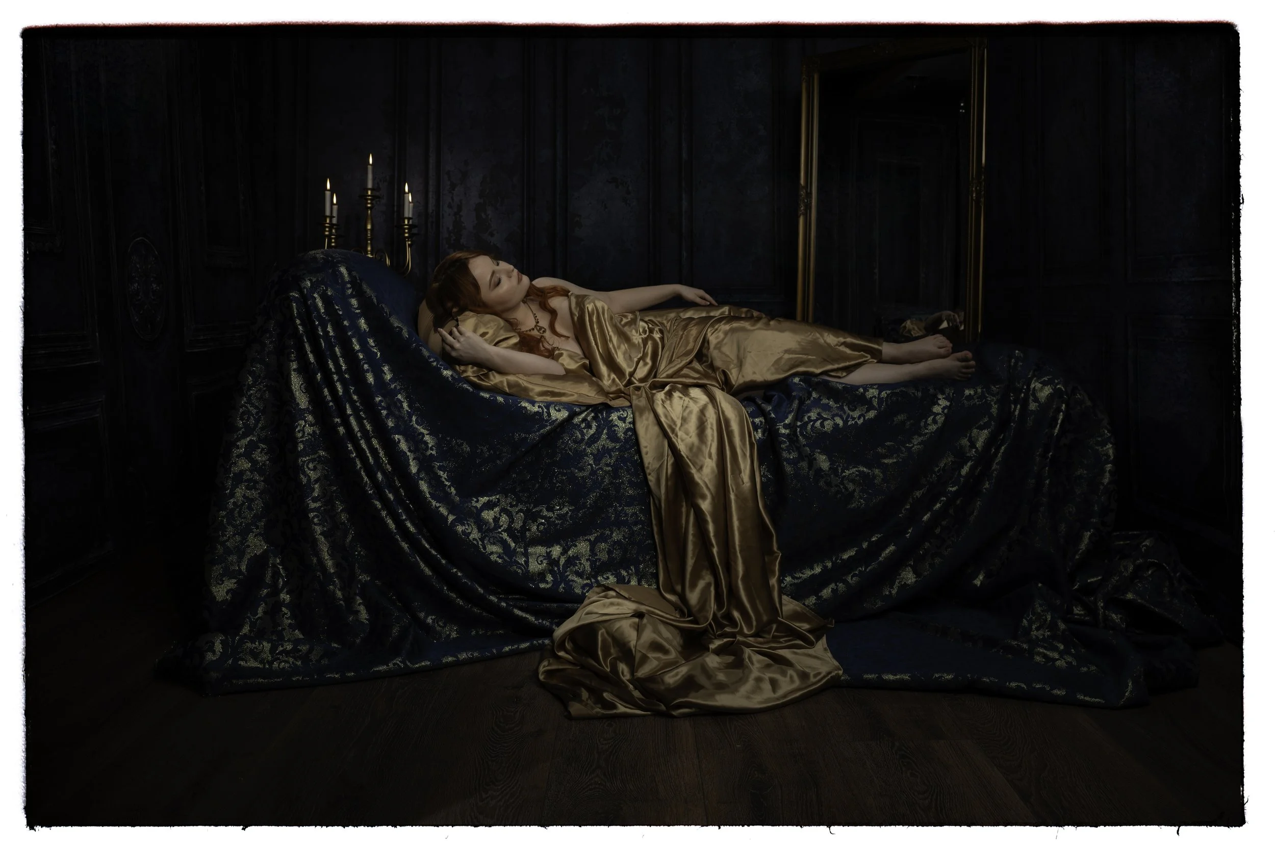 Eine Frau liegt entspannt auf einem Bett in einem dunklen, eleganten Raum, gekleidet in goldene Seide, umgeben von dunkelblauen Vorhängen und Kerzenlicht.