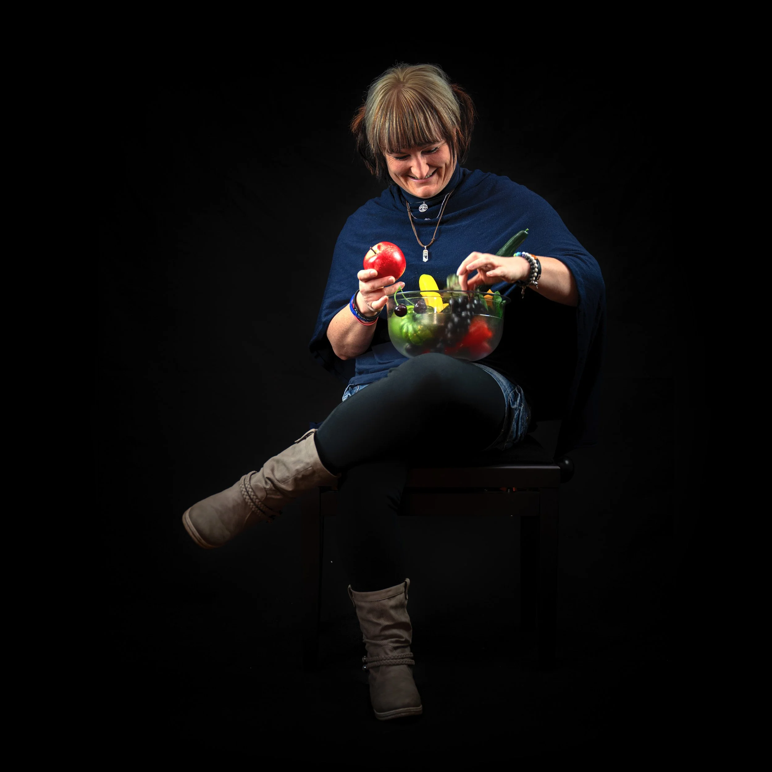 Eine lachende Frau sitzt auf einem Stuhl vor einem schwarzen Hintergrund, hält einen Apfel in der rechten Hand und einen großen Salat mit verschiedenen frischen Früchten und Gemüse in der linken Hand.