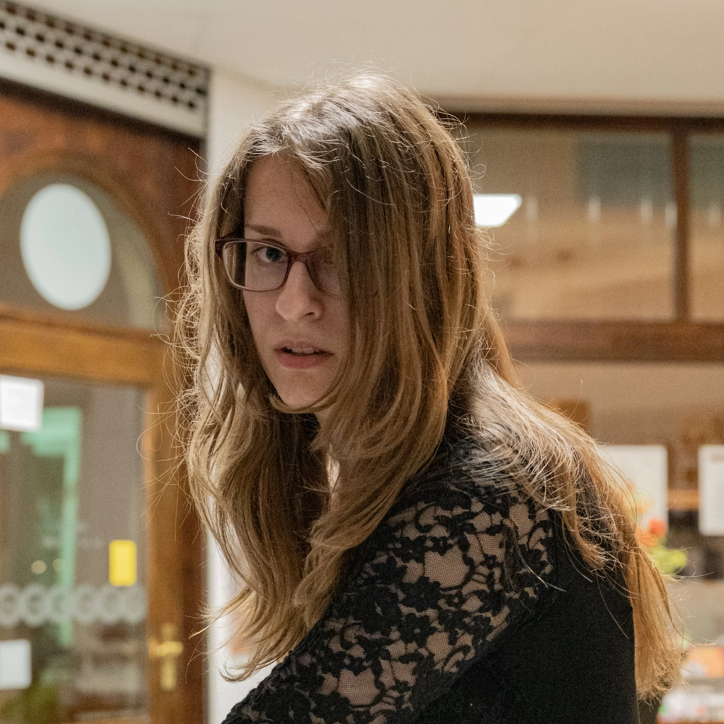 Junge Frau mit langem braunem Haar und Brille trägt schwarzes Spitzenkleid in einem Innenraum.