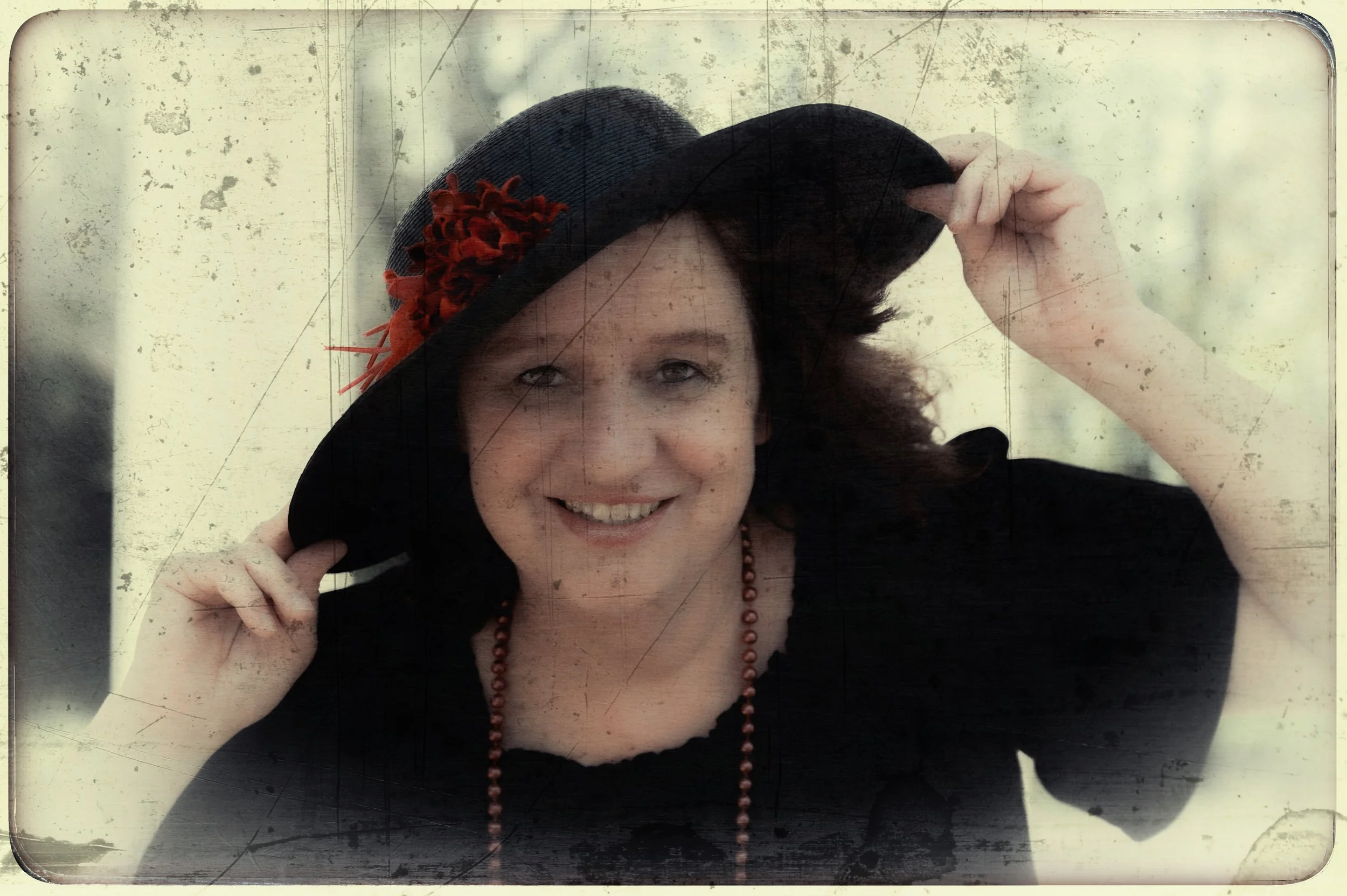 Eine lachende Frau trägt einen schwarzen Hut mit roten Dekorationen, hält den Hut mit einer Hand, trägt eine schwarze Kleidung und eine braune Perlenkette, vor einem hellen Hintergrund.
