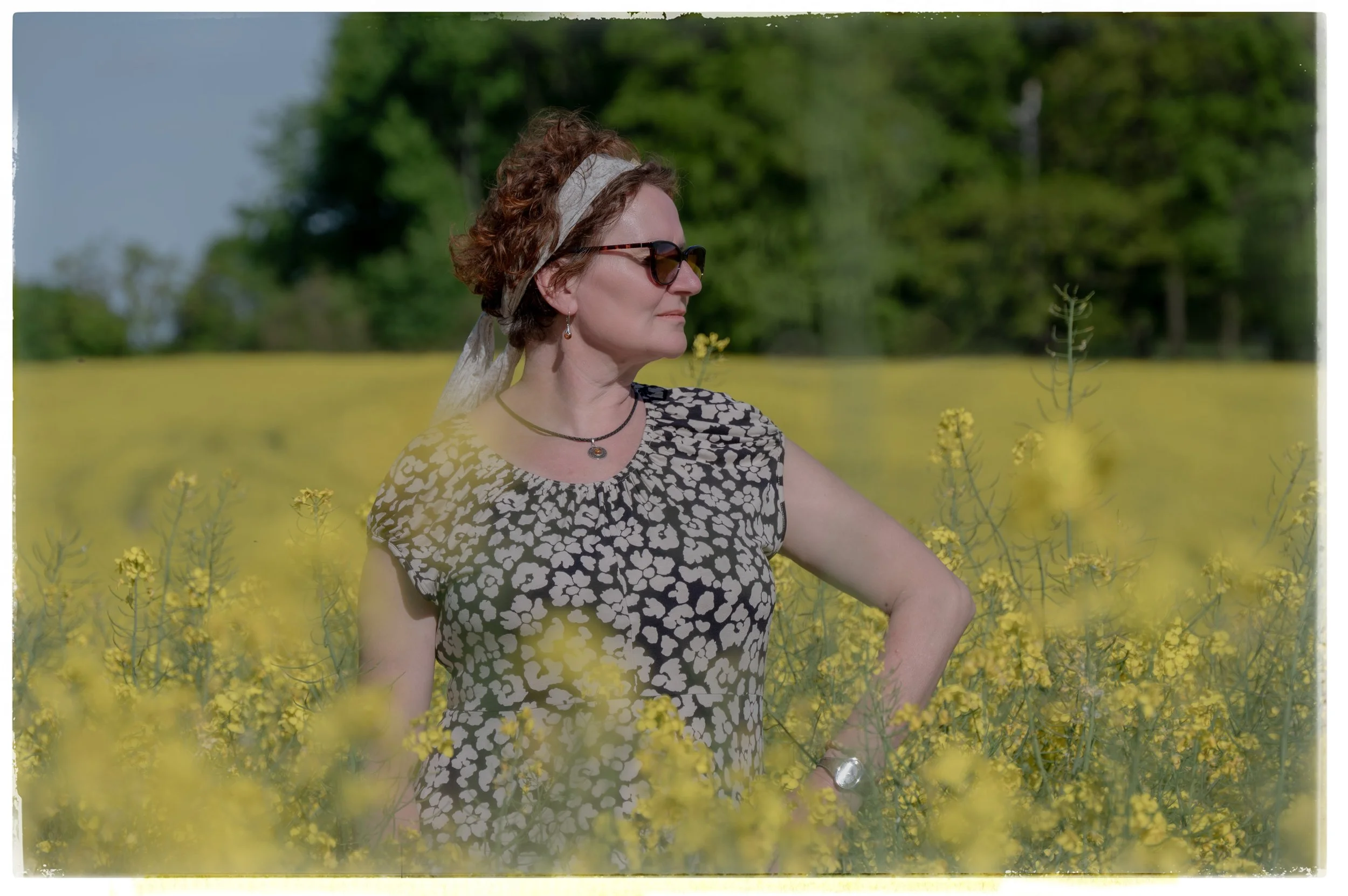 Eine Frau mit braunen, lockigen Haaren, Sonnenbrille und einem grau-weißen Kleid steht in einem gelben Blumenfeld, während sie nach rechts blickt.