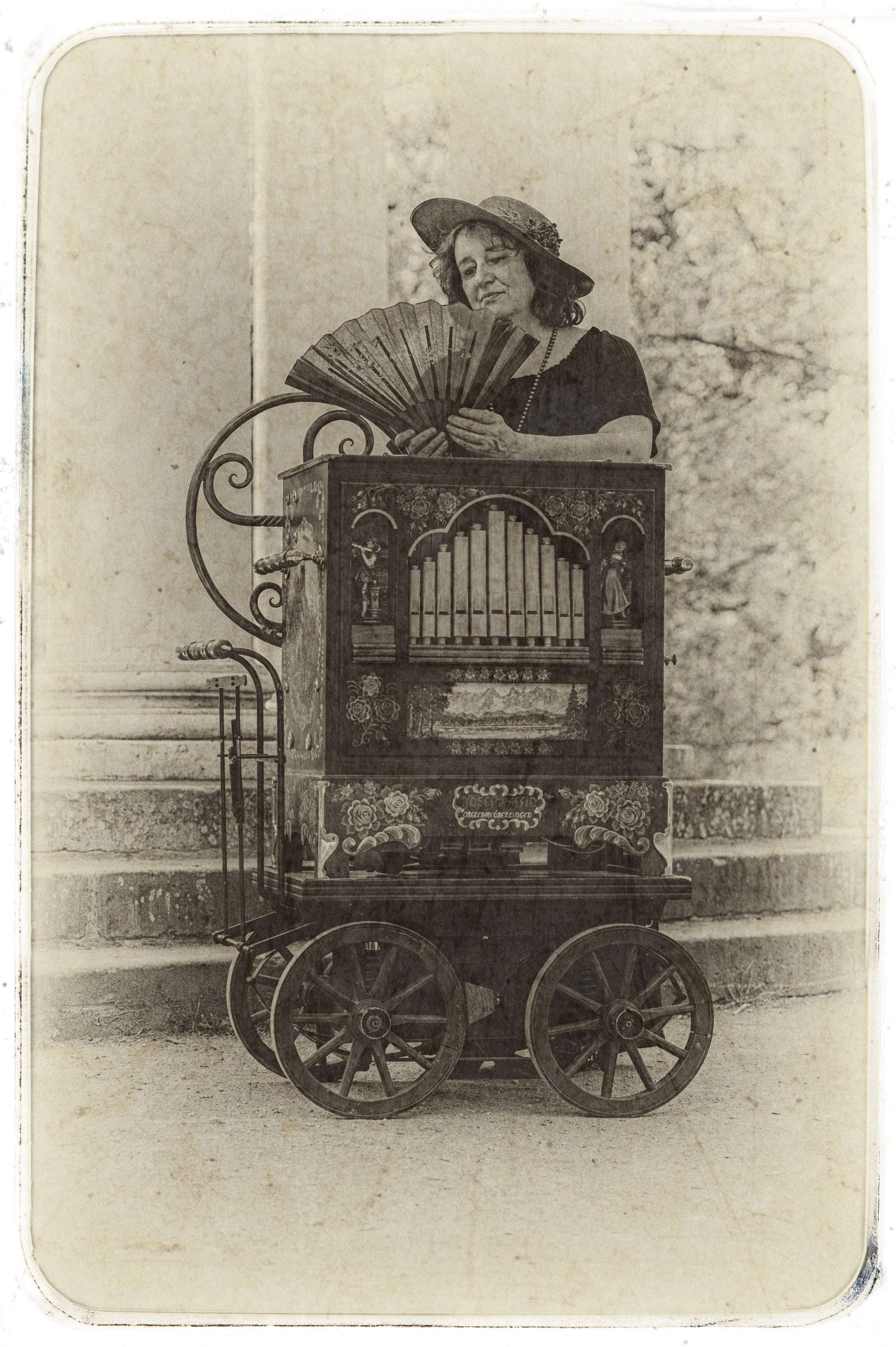 Eine Frau mit Hut steht neben einer historischen Orgel auf vier Rädern, hält einen Fächer und scheint sie zu betrachten.