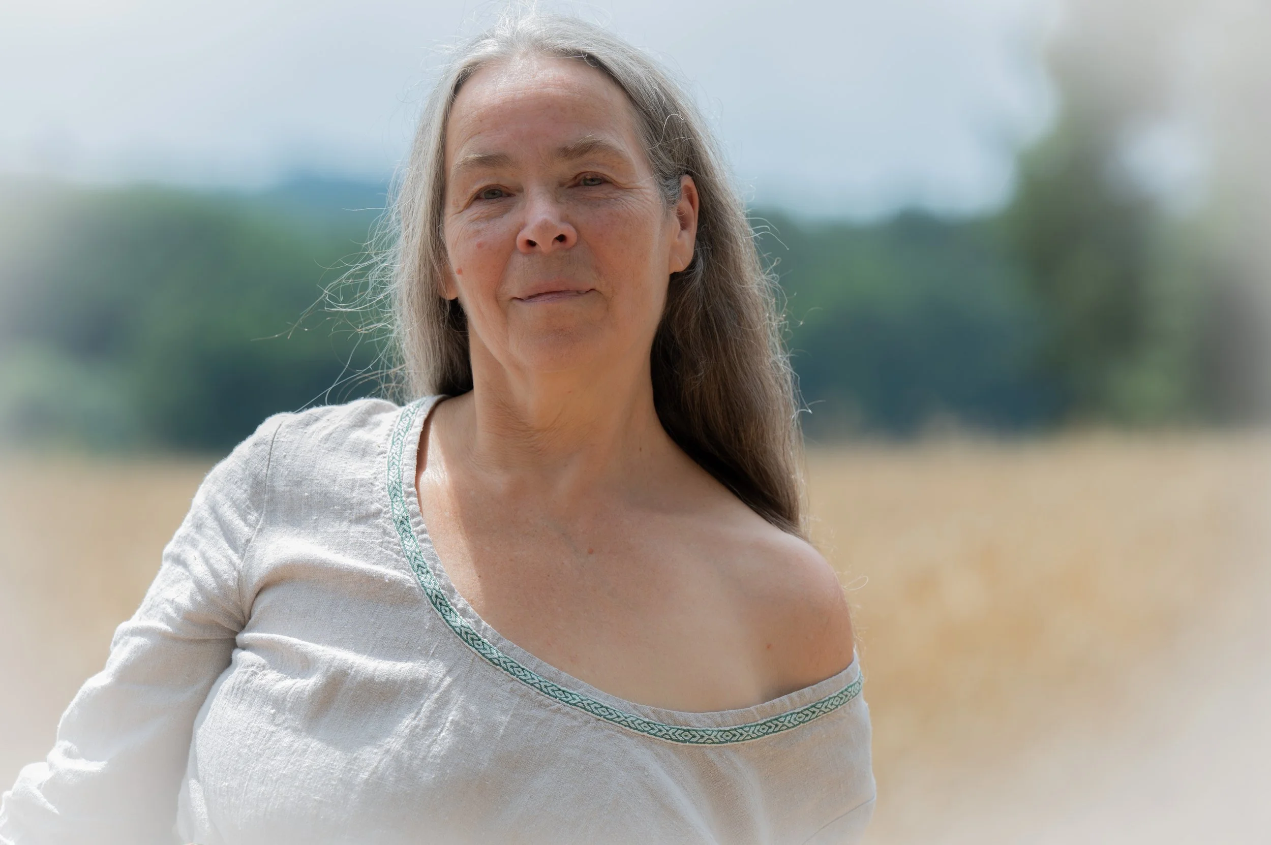 Ein älterer Frau mit langen grauen Haaren steht im Freien, im Hintergrund sind Bäume und ein Feld sichtbar.