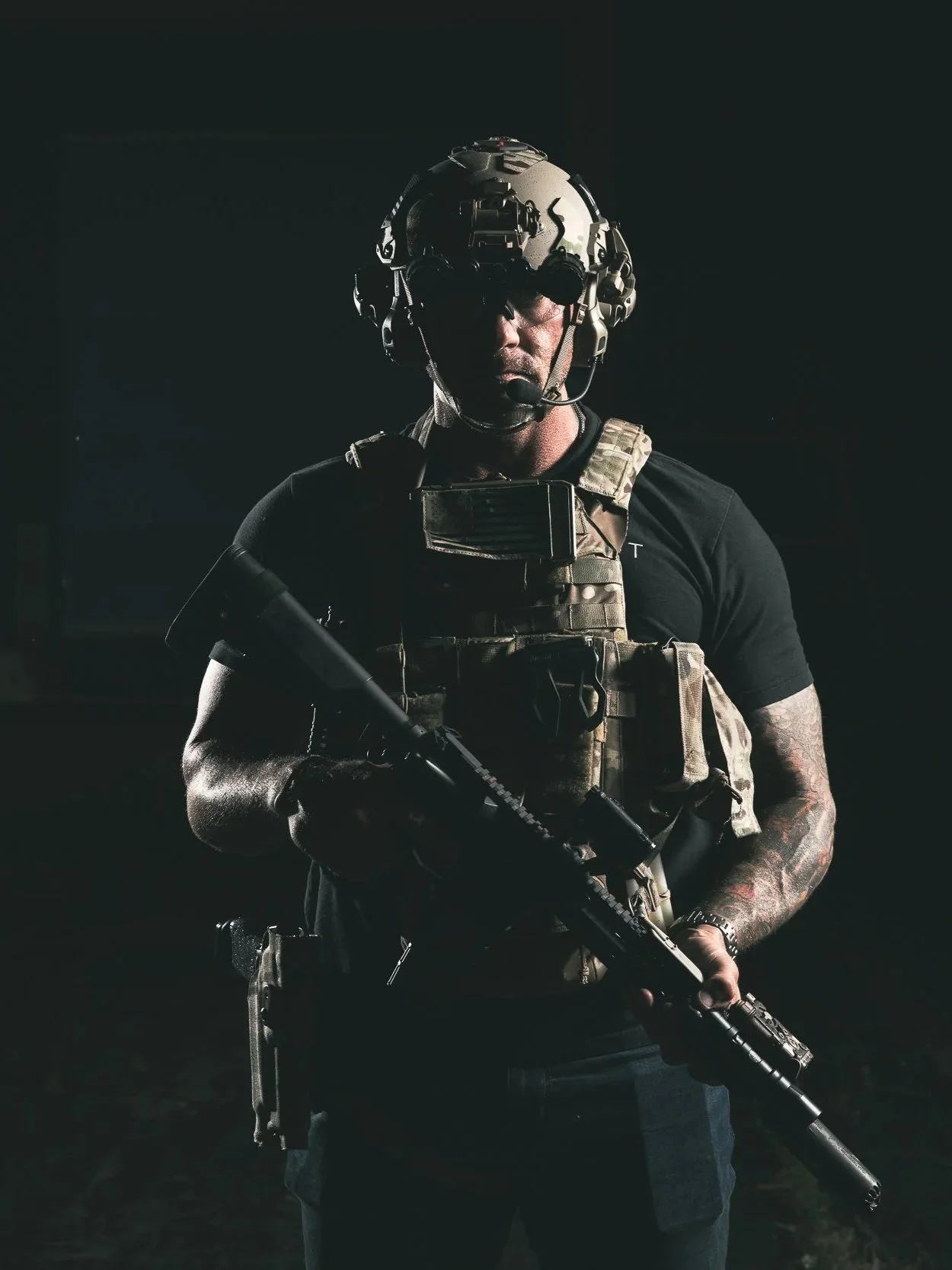 A soldier in tactical gear holding a rifle, wearing a helmet with night vision goggles, and standing in a dark environment.