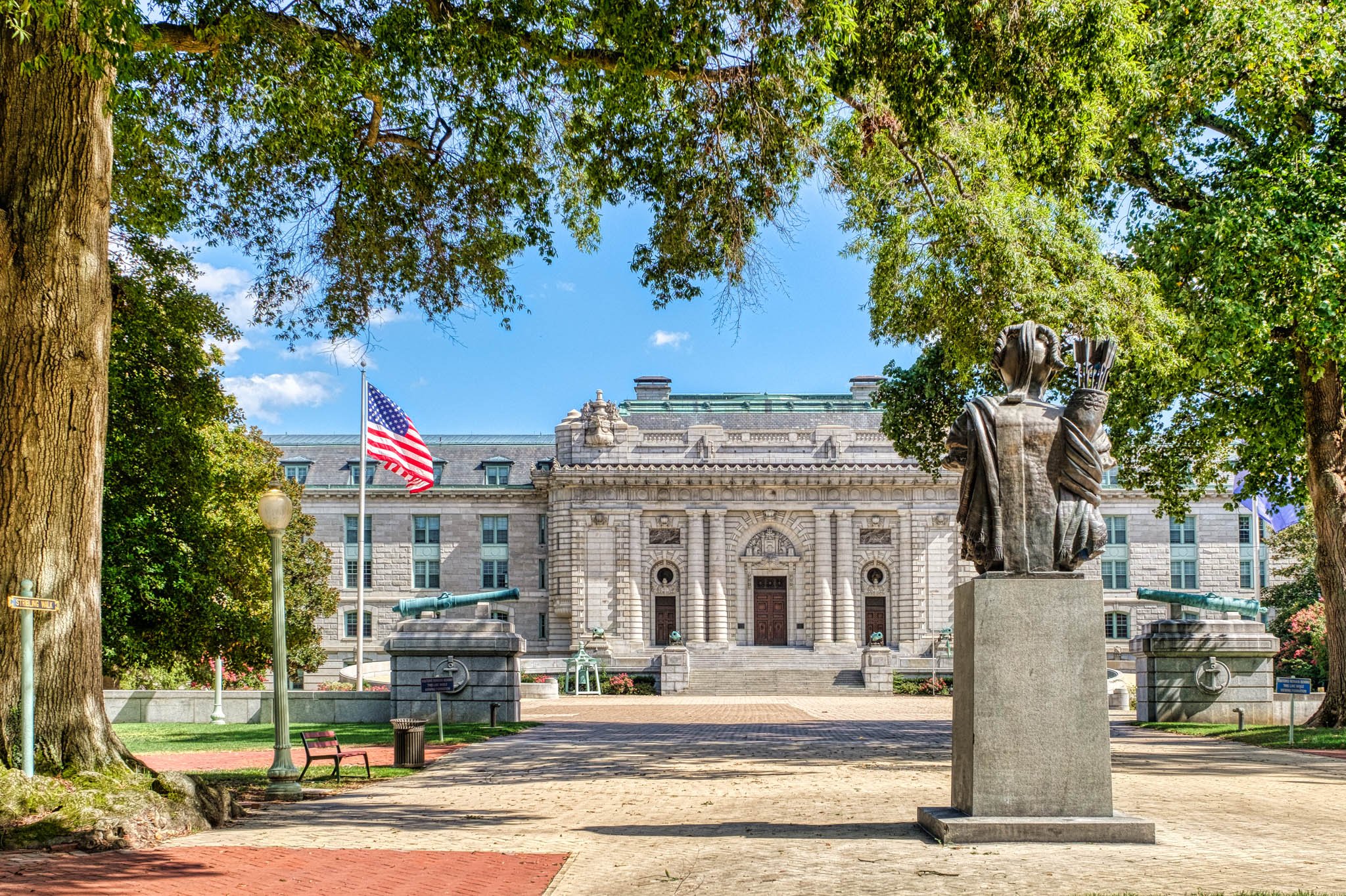 A historic building with columns and a dome, flanked by trees, with a statue of a woman holding a quill and scroll in the foreground.