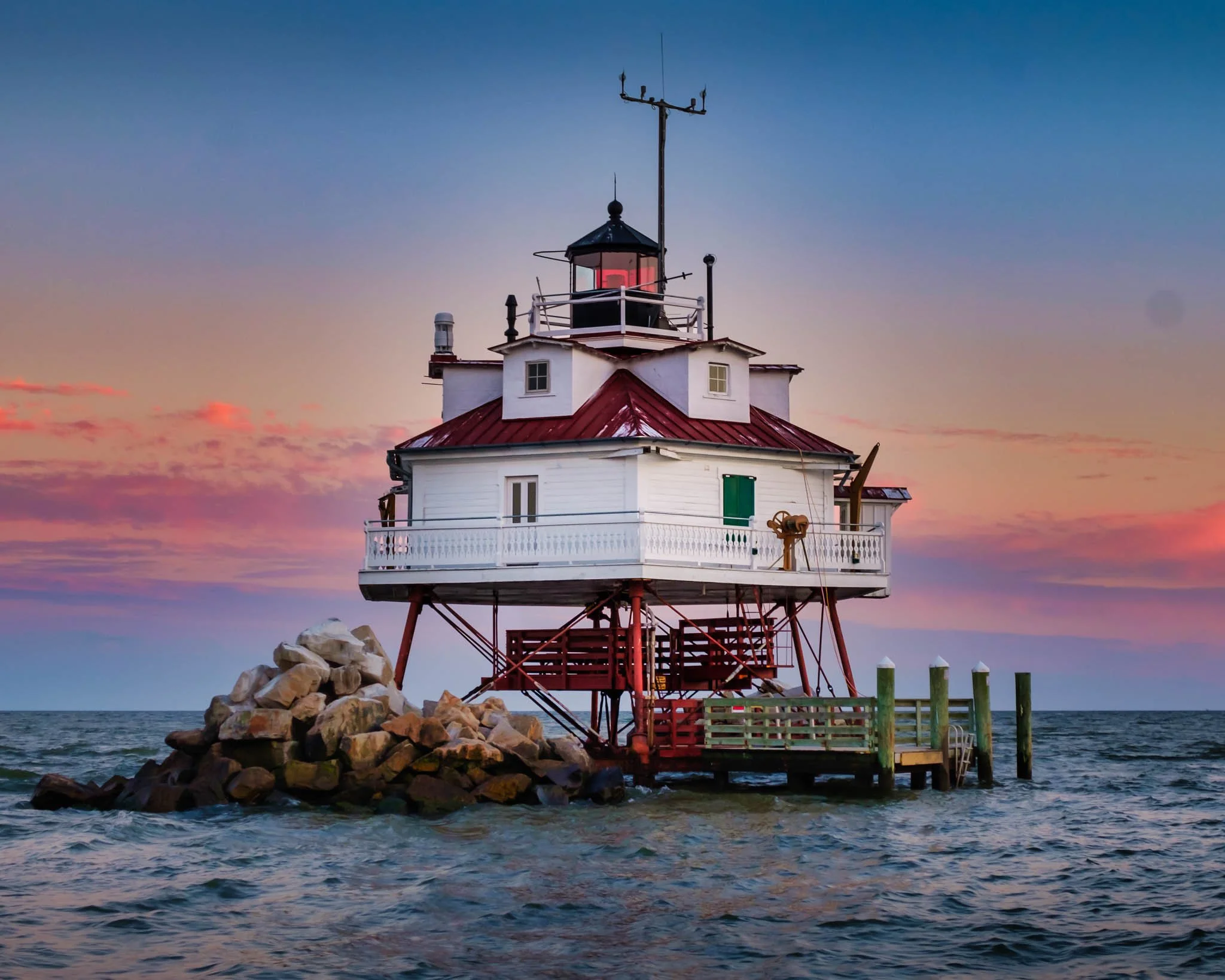 A lighthouse with a red roof and white walls on a rocky base in the water during sunset with colorful sky.