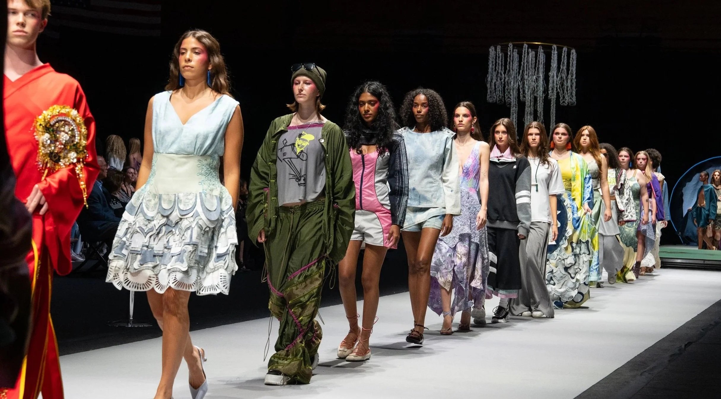 Models walk down a runway during a fashion show, showcasing diverse and eclectic outfits in a dark venue with an audience watching.