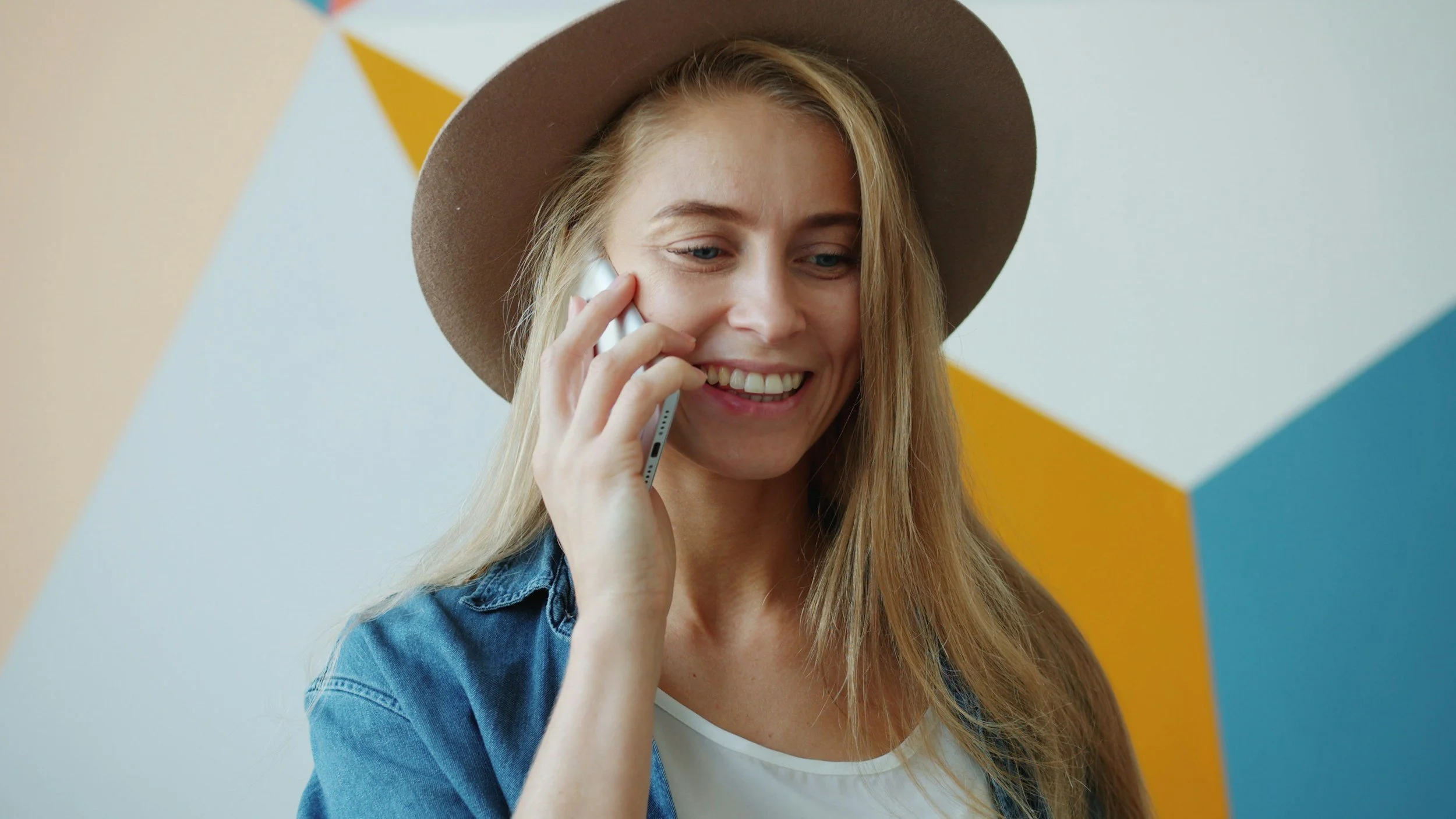 A young woman with long blonde hair, wearing a tan hat and denim jacket, is talking on her cellphone and smiling. The background features a modern, geometric wall with white, blue, and yellow shapes.