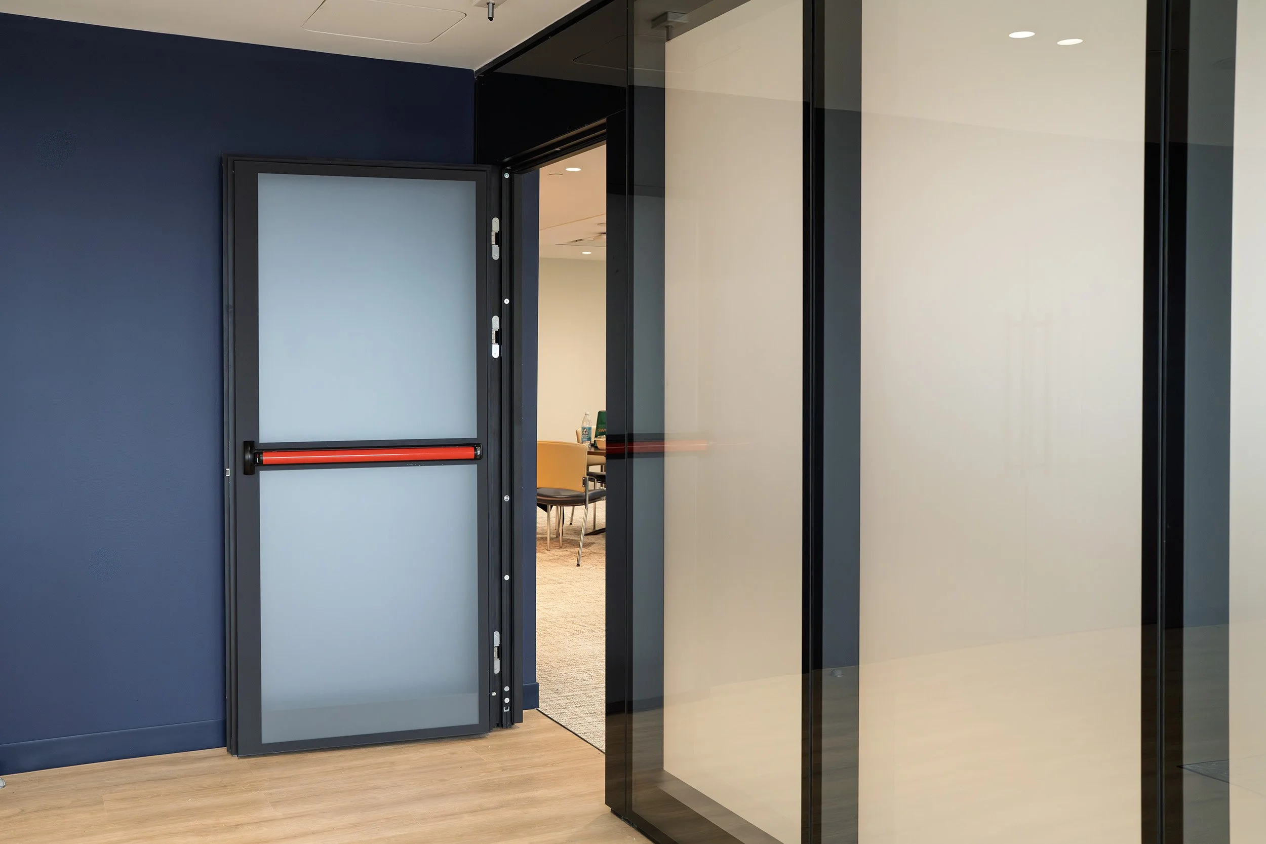 An interior view of a modern room with a glass door and a blue wall. The door has a red push bar and is partially open, revealing a meeting room with chairs and a water bottle on a table.