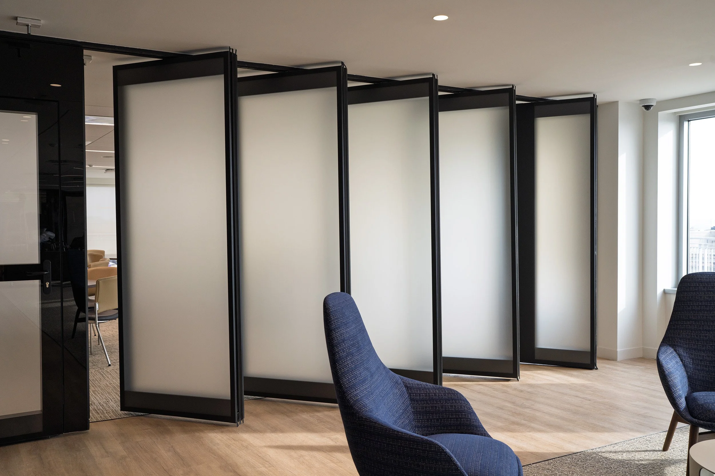 Office space with black-framed white room dividers, blue chairs, a window, and a glass door.
