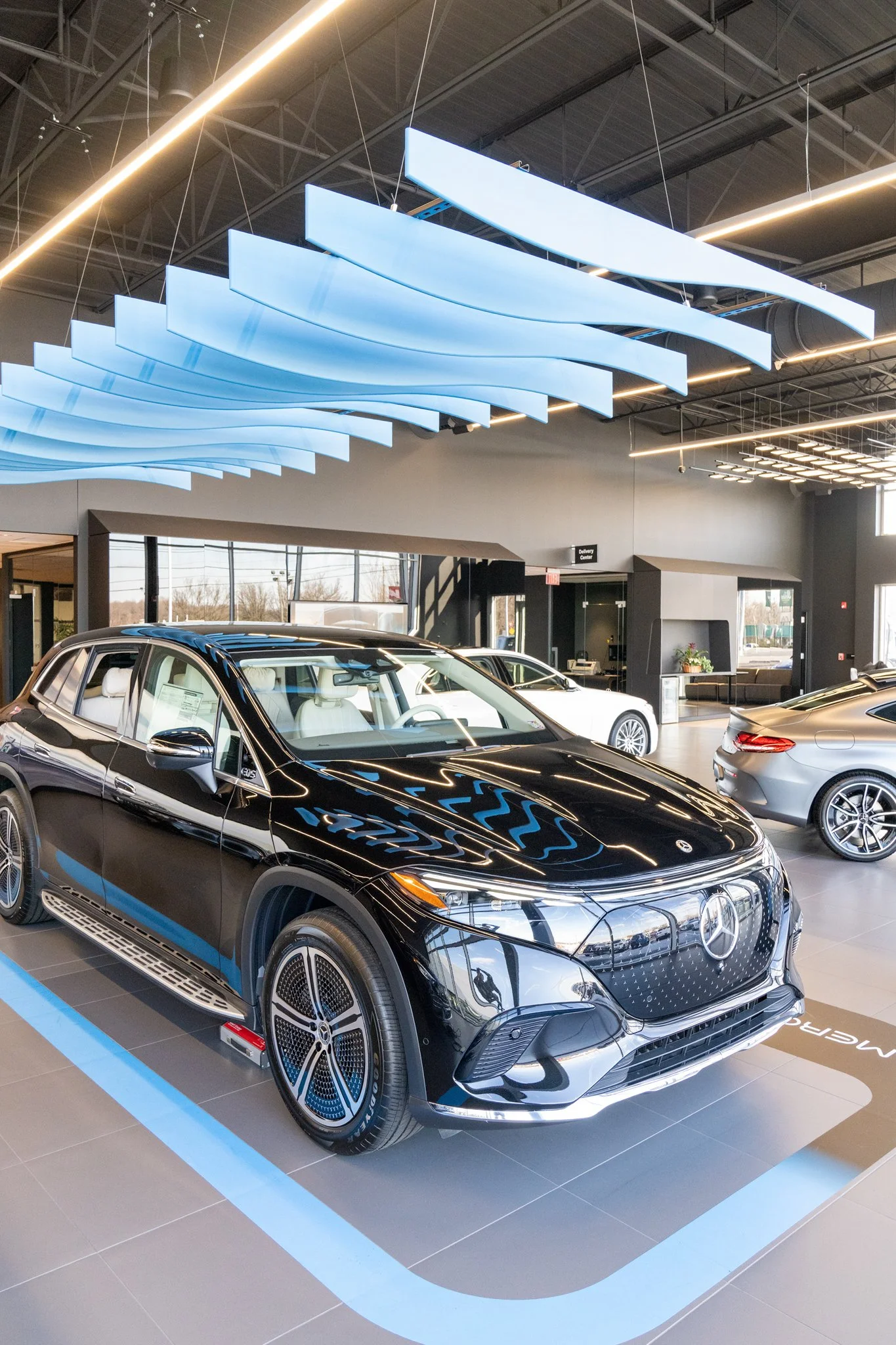 Interior of a car dealership showroom displaying a black Mercedes-Benz electric vehicle with white interior, and several other cars in the background.