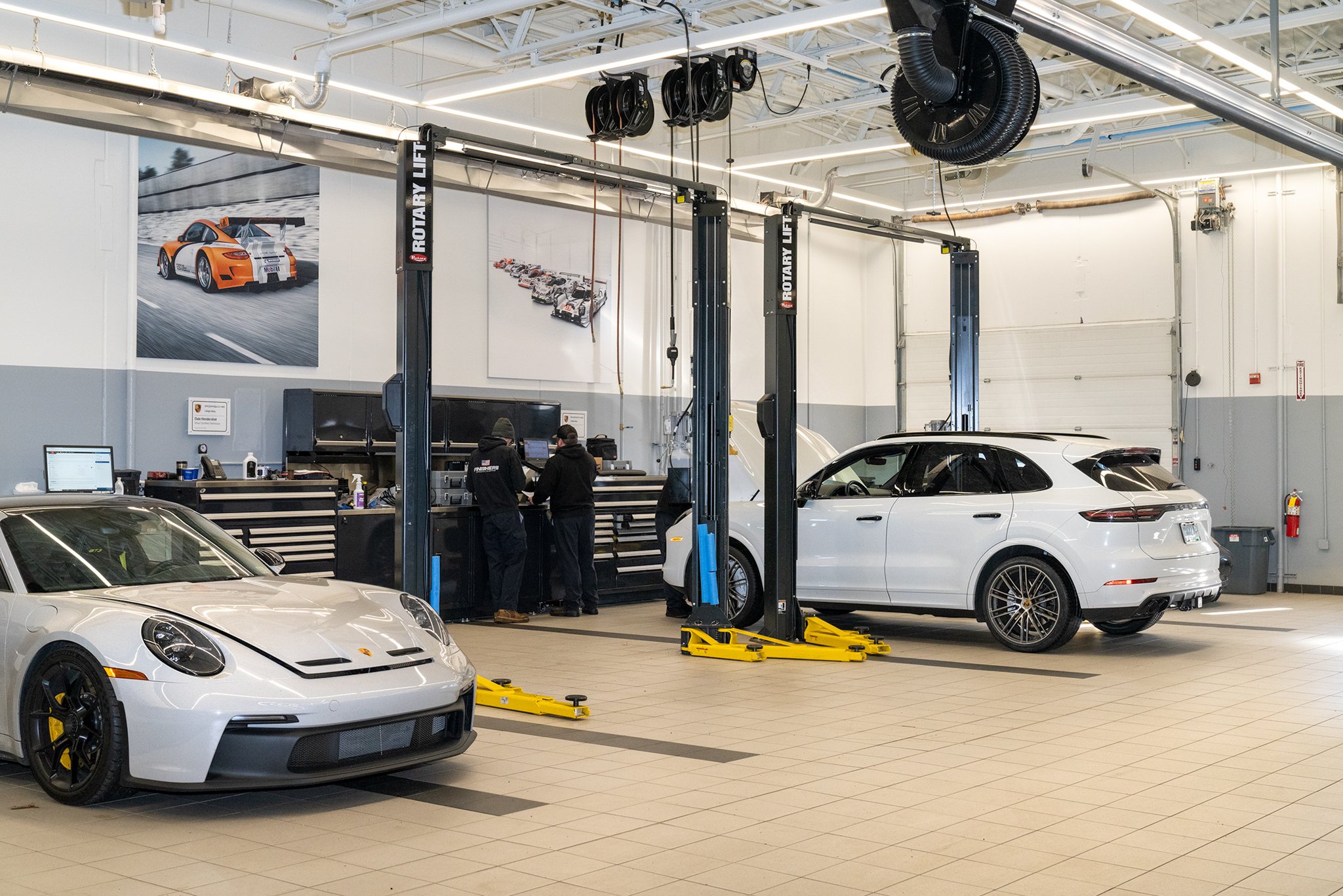 Inside an automotive repair shop with two white luxury cars, a Porsche and a Genesis SUV, on hydraulic lifts, and a silver Porsche 911 parked on the floor. There are three workers at a workstation with tools, computers, and equipment. The shop has a 