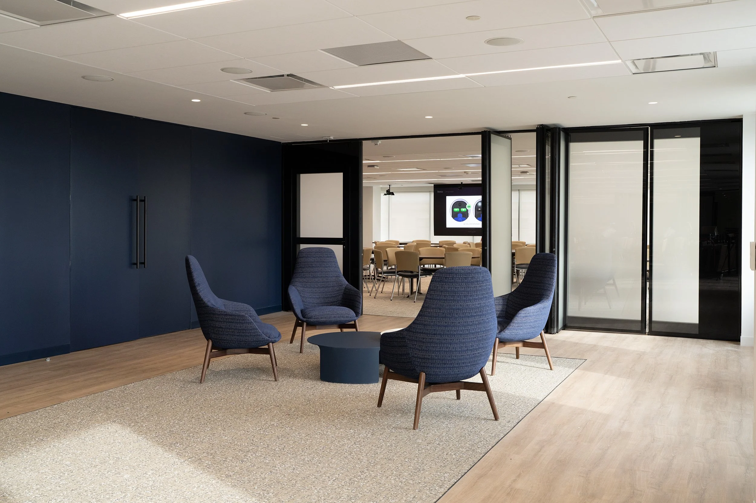 Modern office lounge area with four navy blue armchairs surrounding a small round table, beige carpeting, and glass partitions leading to a conference room with beige chairs and two screens.