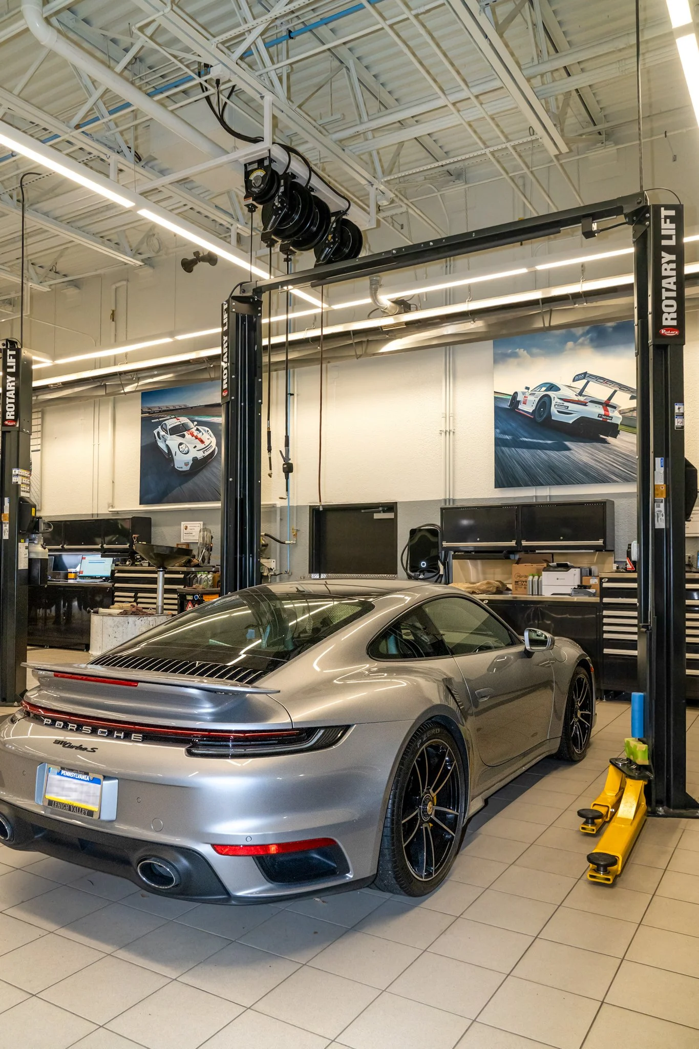 A silver Porsche sports car inside an automotive workshop with a hydraulic lift system. The workshop has two framed photos of racing cars on the wall and various tools and equipment on black tool cabinets.